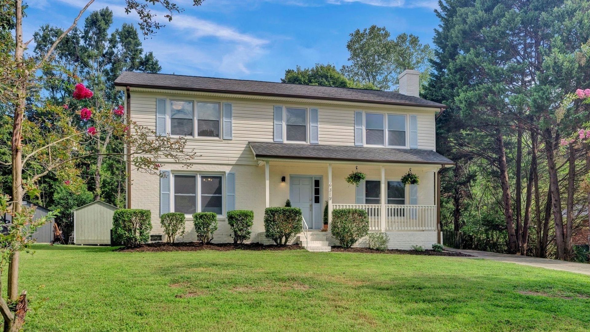 Two-story beige house with light blue shutters, front porch with hanging plants, surrounded by green lawn, bushes, pink flowers on left, and tall trees on right under a blue sky.