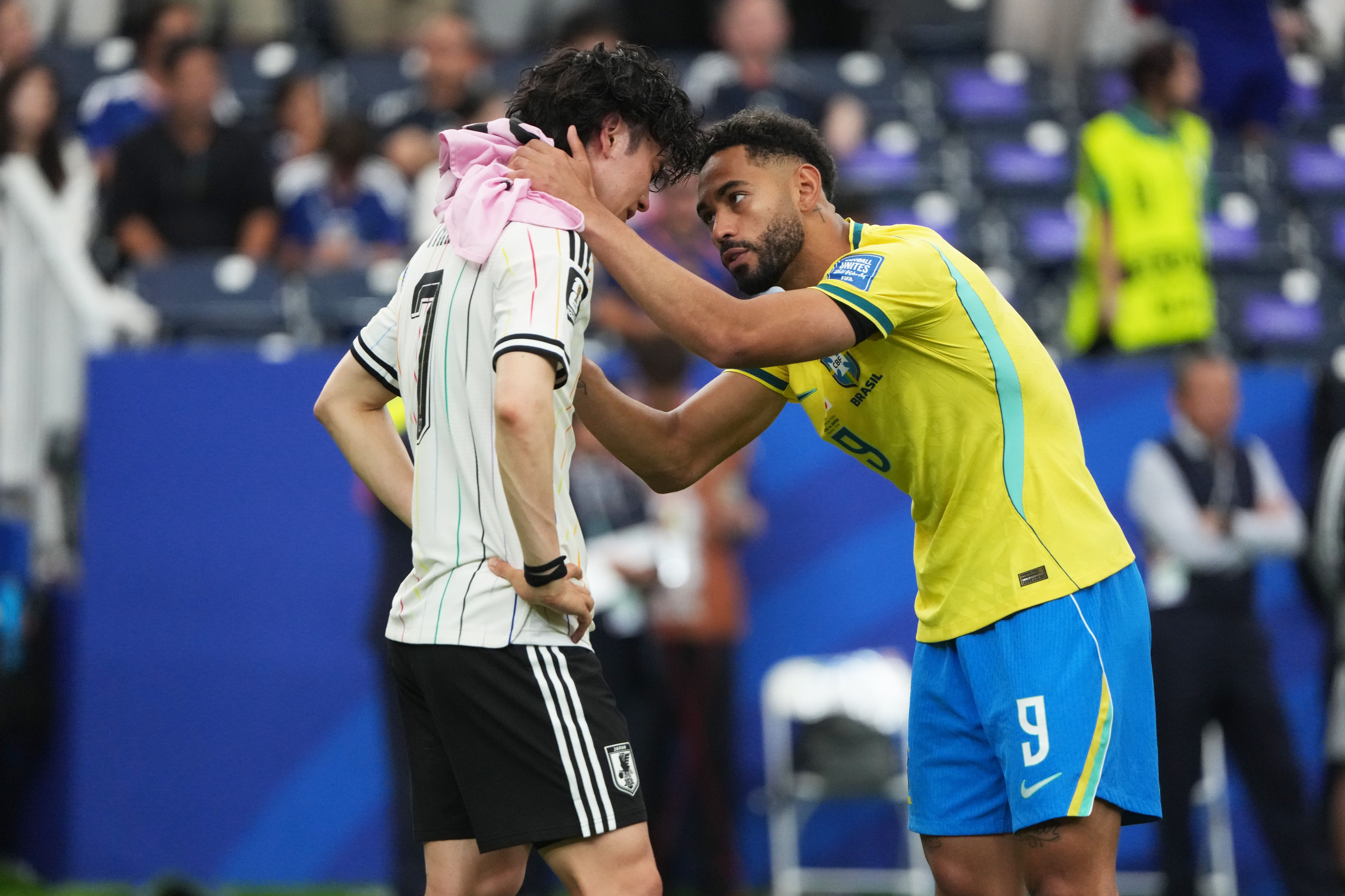 Two male soccer players face off in the foreground: one in a white jersey with a pink hoodie, the other in a yellow Brazil kit with blue shorts; their foreheads touch as hands grip.