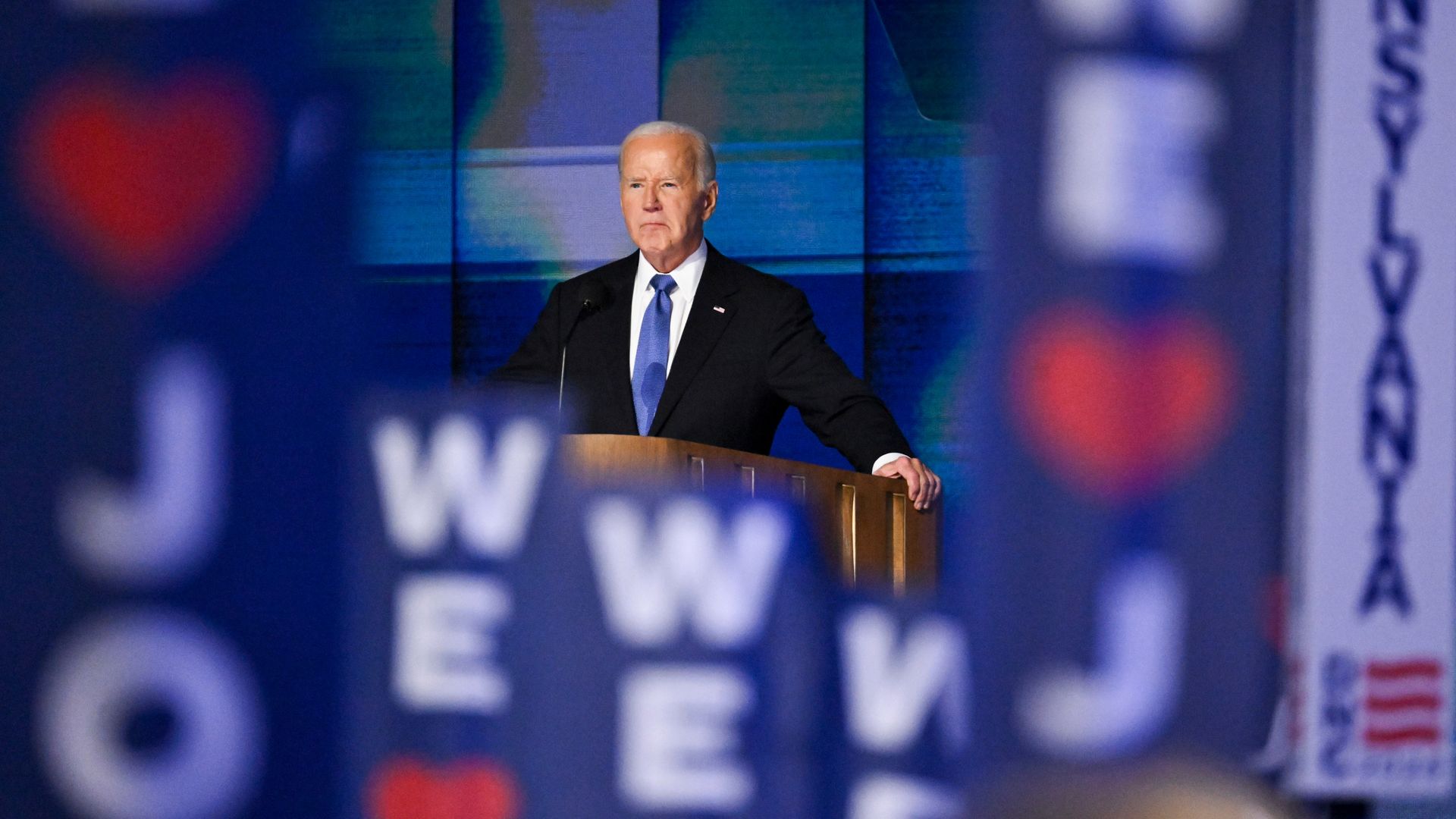 President Biden, wearing a dark gray suit and gripping a wooden podium in front of a blue backdrop with verticle "We <3 Joe" sign sticks in the foreground. 
