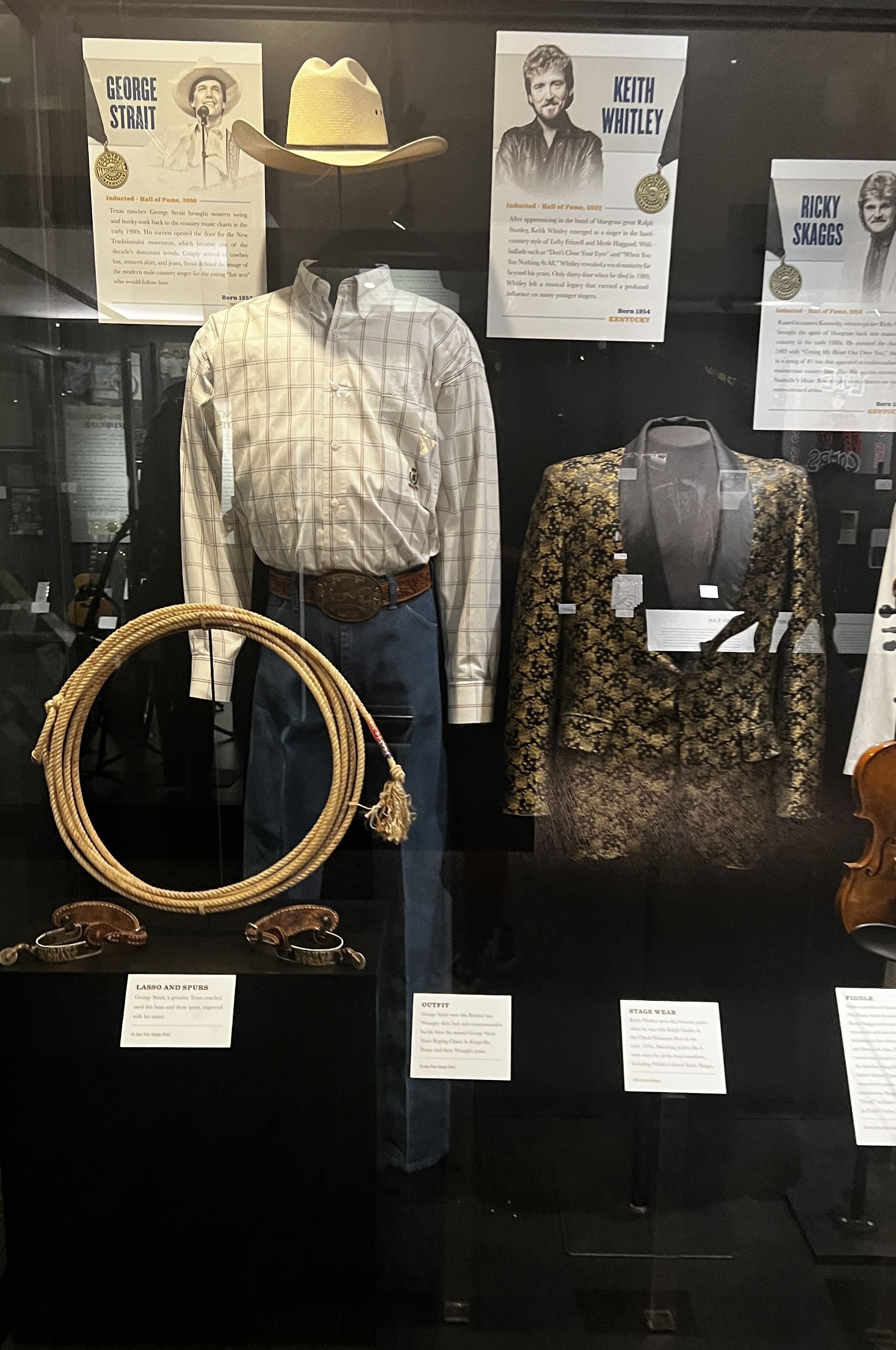 A cowboy outfit, lasso and suit behind a glass case. 