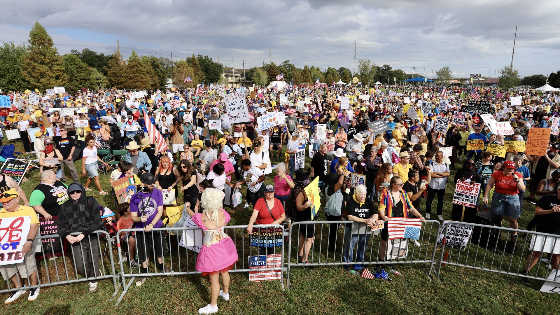 Large crowd gathered outdoors for a protest with various colorful signs and American flags, many wearing hats and sunglasses, under a partly cloudy sky.