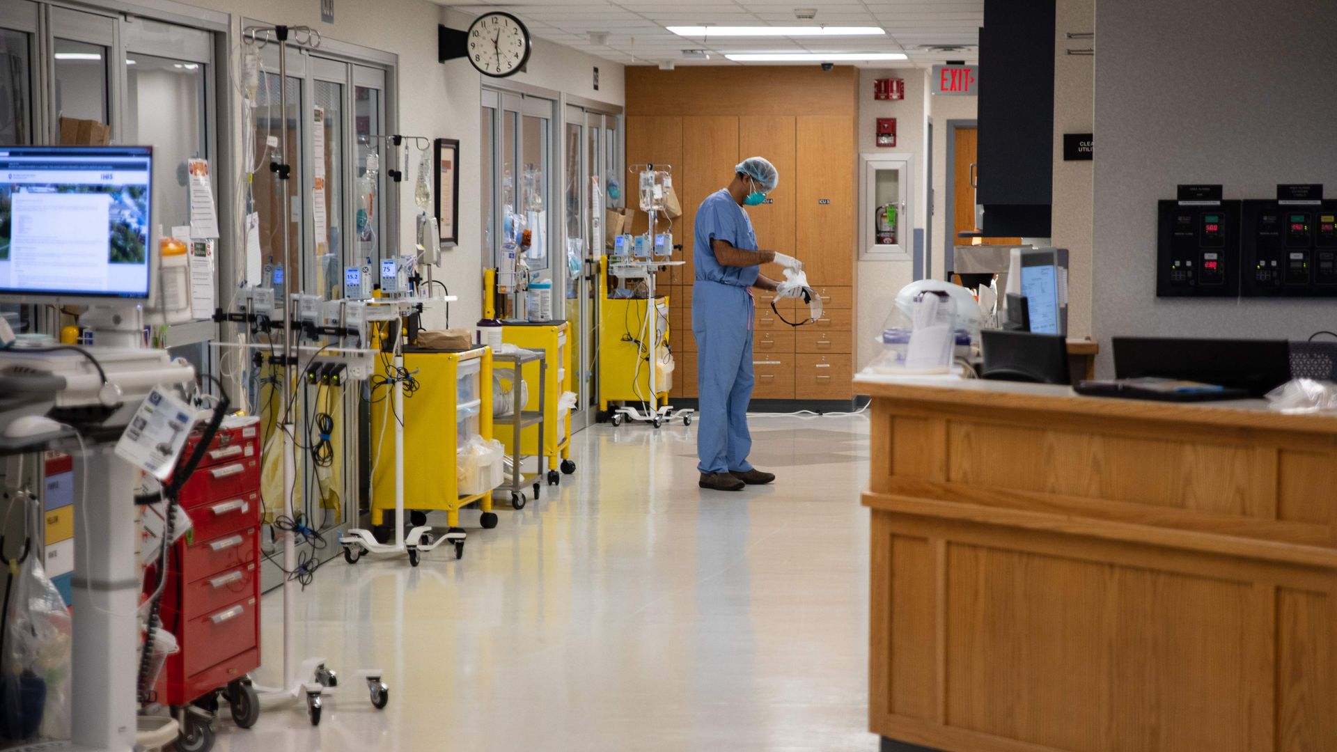 A doctor in blue protective gear stands in a hospital COVID-19 unit with medical equipment outside of rooms.