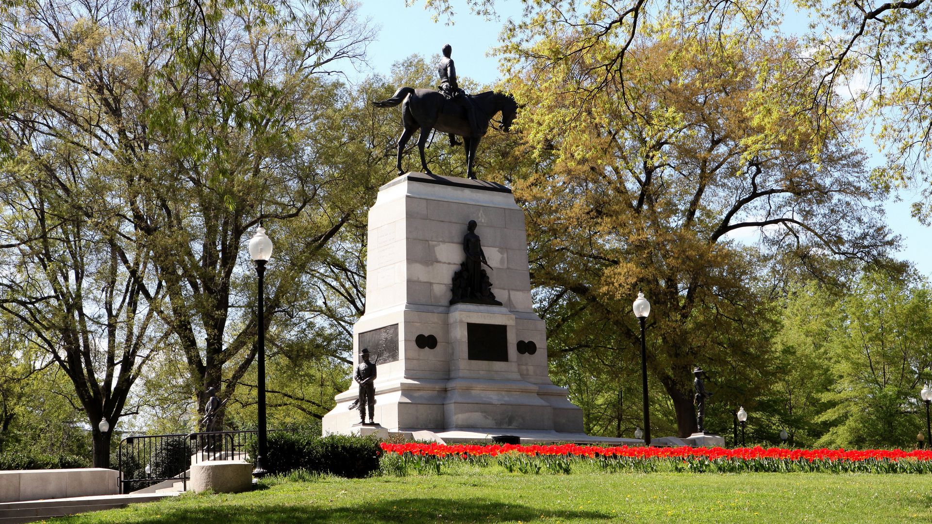 Equestrian statue atop a tall stone pedestal in Sherman Park, rider on a horse overlooking a bed of bright red tulips. Surrounding trees and vintage lampposts frame the monument.