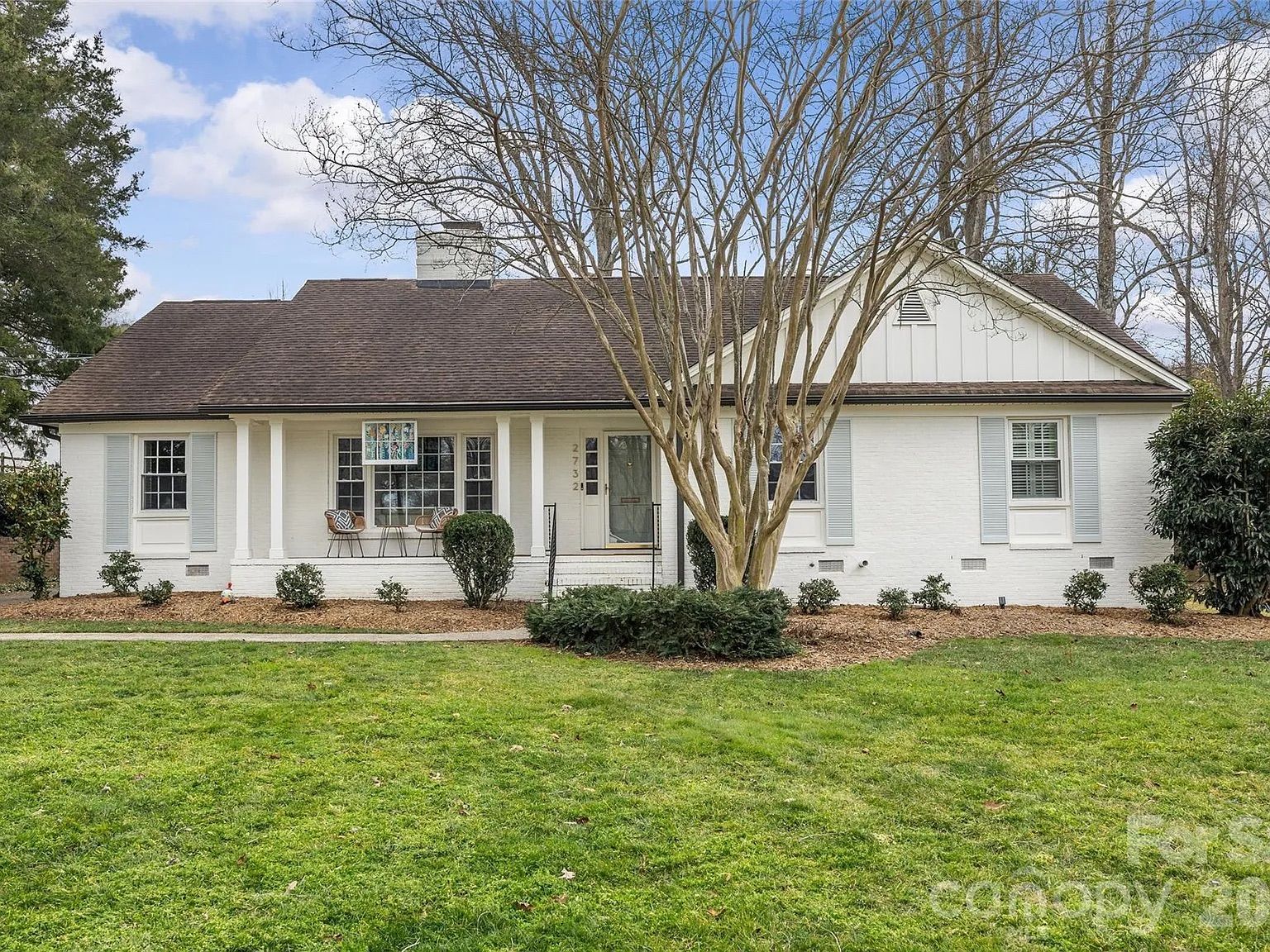 White brick ranch-style house with a brown shingle roof, a front porch with two chairs, a leafless tree in the yard, trimmed shrubs, and a green lawn under a blue sky.