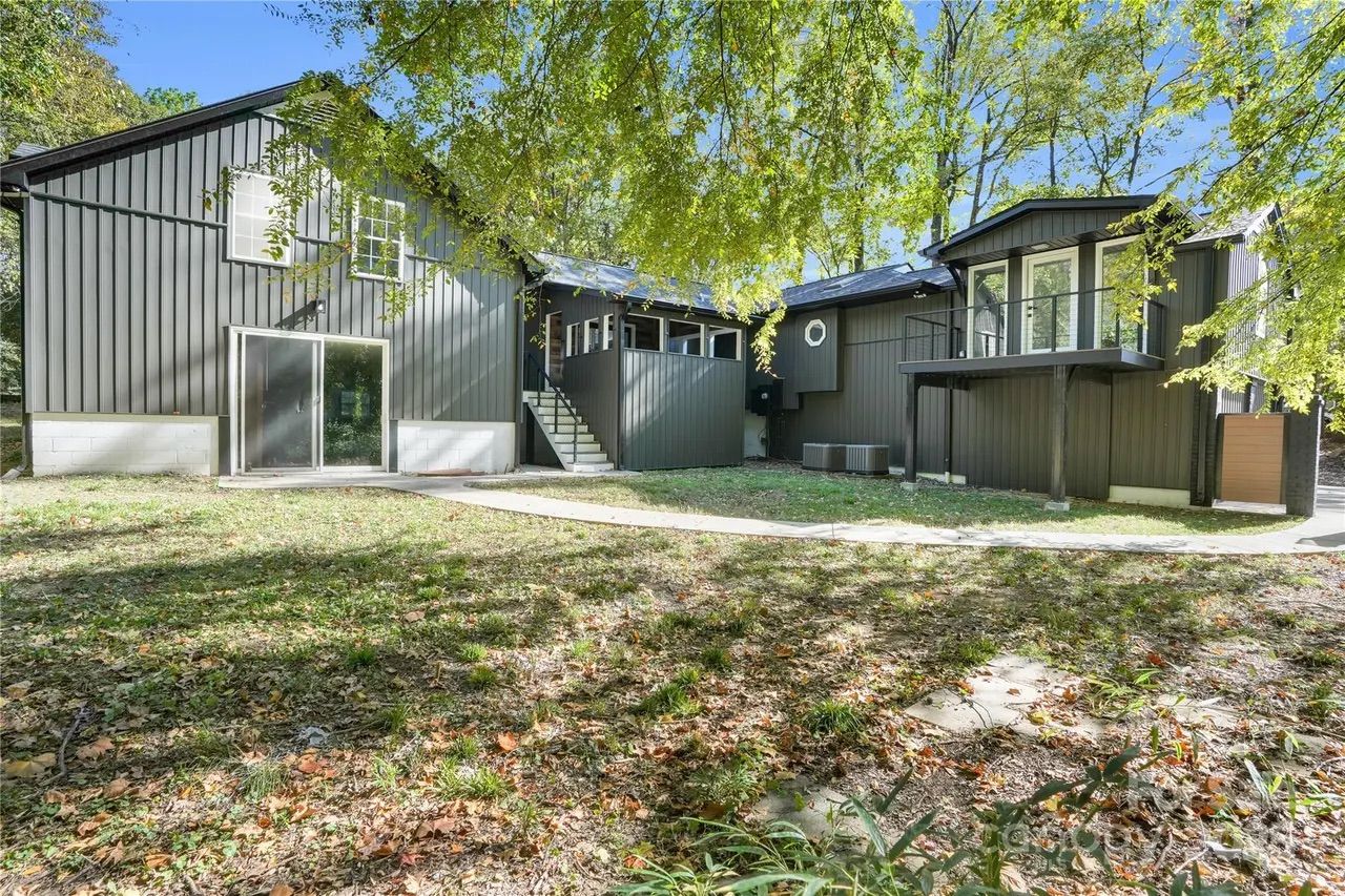 Gray modern house with vertical siding, large windows, sliding glass door, and balcony, surrounded by green trees and a lawn with scattered leaves under a clear blue sky.