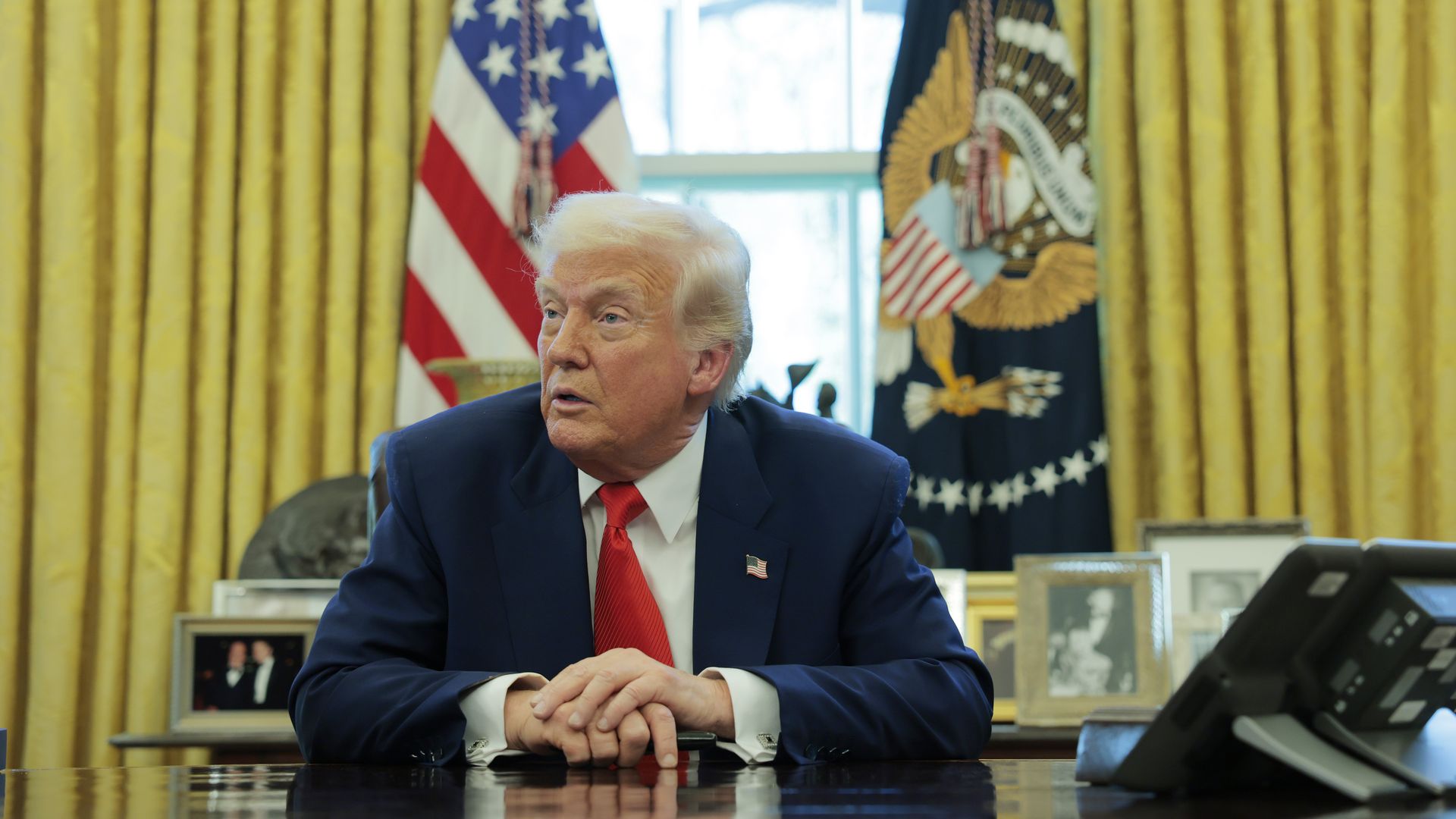 President Trump wearing a blue suit and putting his hands on the resolute desk in front of the White House's gold curtains.