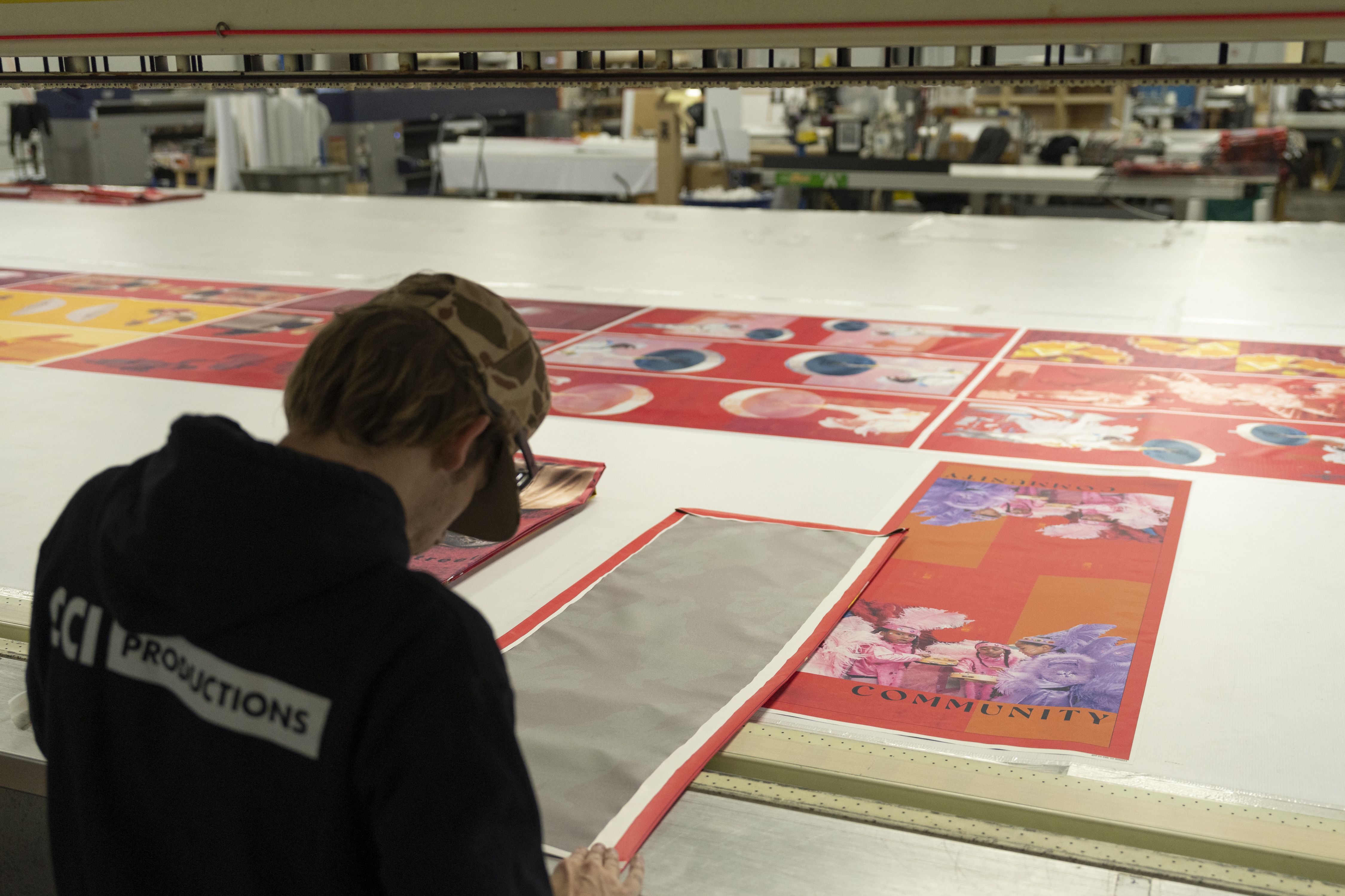 Person wearing camo hat and black hoodie inspecting printed red and orange fabric panels on a large white table in a workshop.