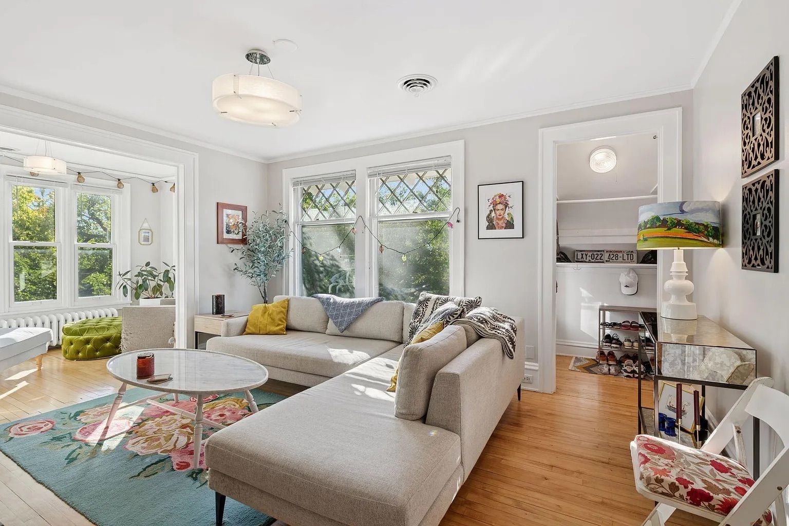 Bright living room with beige L-shaped sofa, floral rug, round white coffee table, large windows, potted plants, colorful cushions, and a lamp with painted lampshade on a mirrored table.
