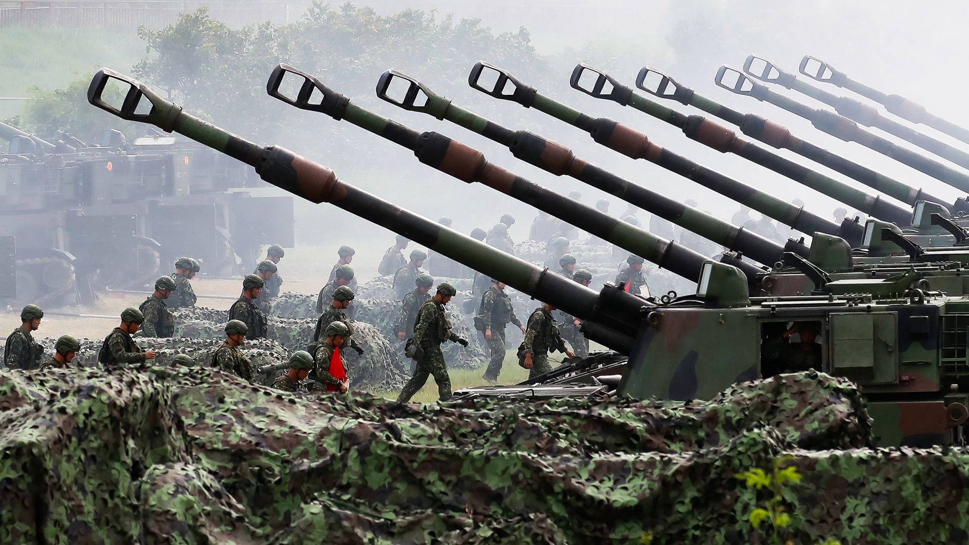 Row of camouflaged self-propelled artillery vehicles with long barrels pointing upward, soldiers in green uniforms and helmets walking alongside on a misty day.