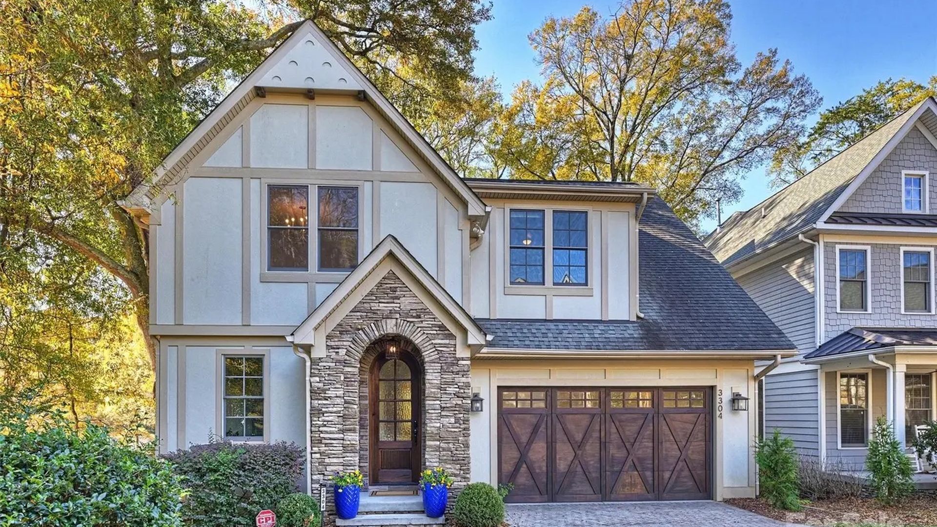 Two-story beige house with stone entrance arch, dark wooden front door, two blue flower pots, and large brown garage door, surrounded by green bushes and trees under a blue sky.