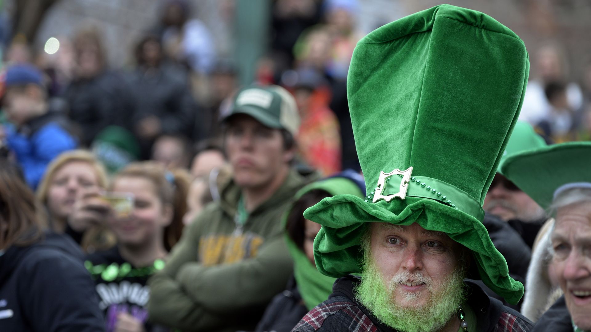 A crowd scene, with a man wearing a ridiculously large green hat with a golden buckle in the foreground. His beard is dyed green.