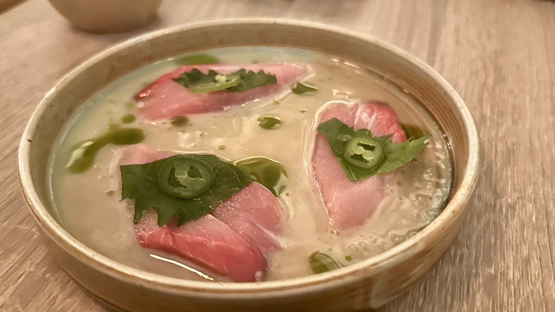 A round ceramic bowl of pale, milky broth with pink sashimi slices, topped with cilantro leaves and thin green chili slices, on a light wooden table.