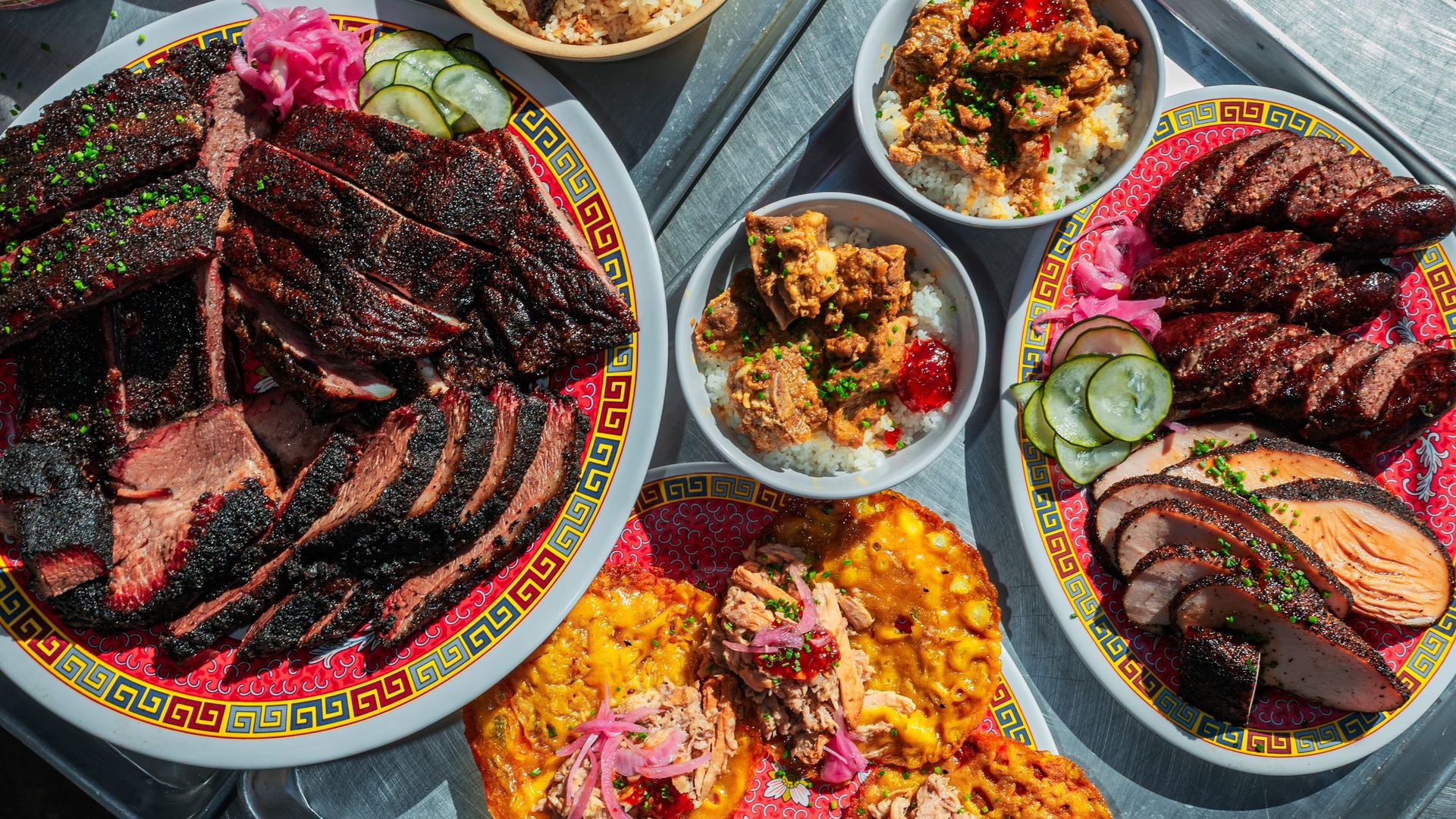 Outdoor metal table feast: multiple bowls of rice topped with char siu pork and greens, plus plates of sliced roast beef and assorted side dishes, all basking in sunlight.