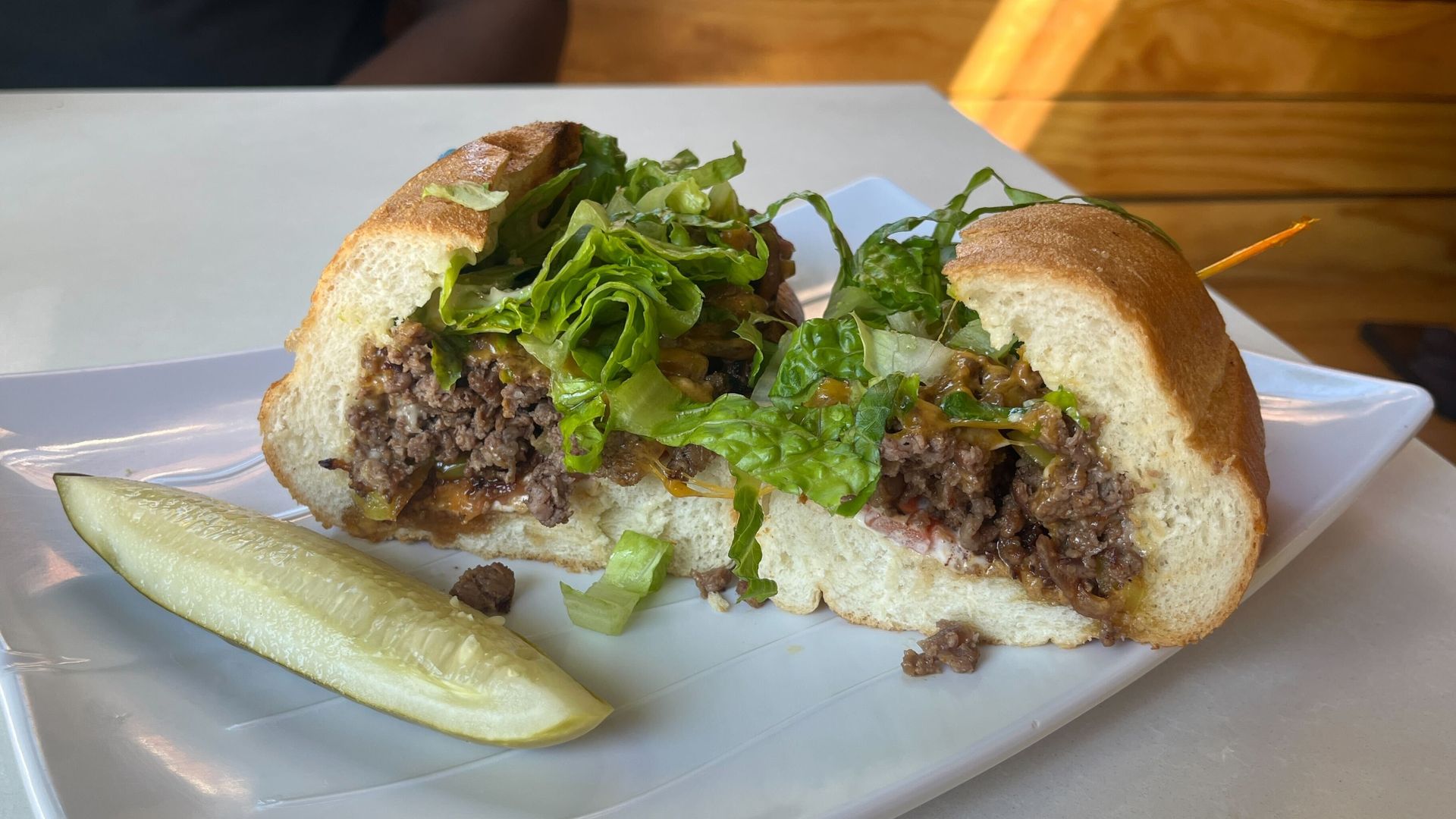 A sandwich cut in half on a white plate, filled with lettuce and ground beef, served with a dill pickle spear on the side.