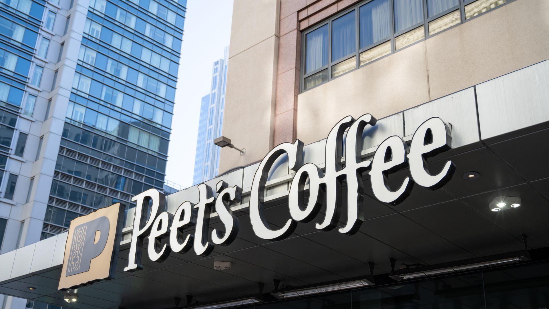 Exterior view of a Peet's Coffee shop sign on a building in an urban area with tall glass skyscrapers in the background under a clear sky.