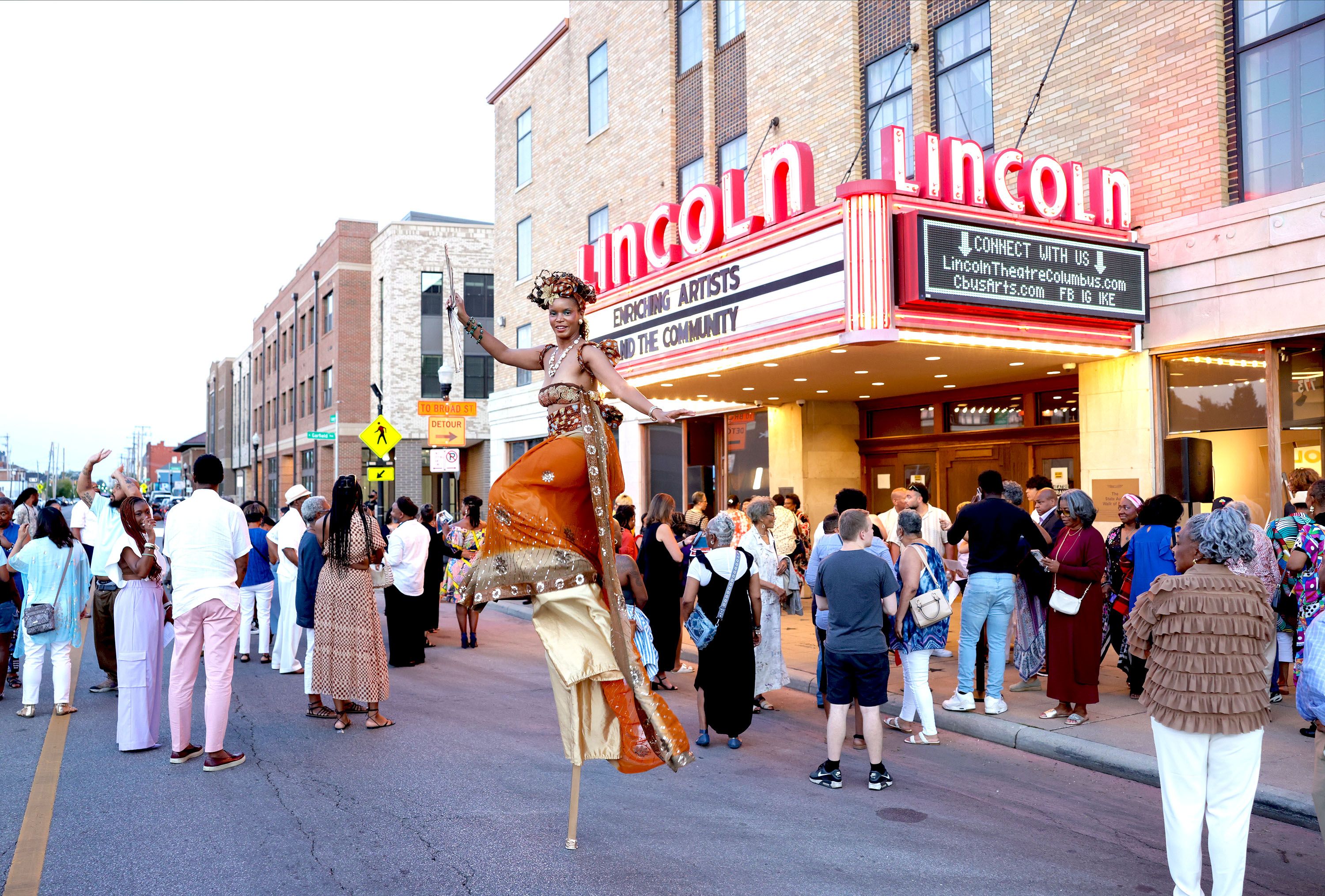 A stilt performer walks amid a crowd in front of the Lincoln Theatre