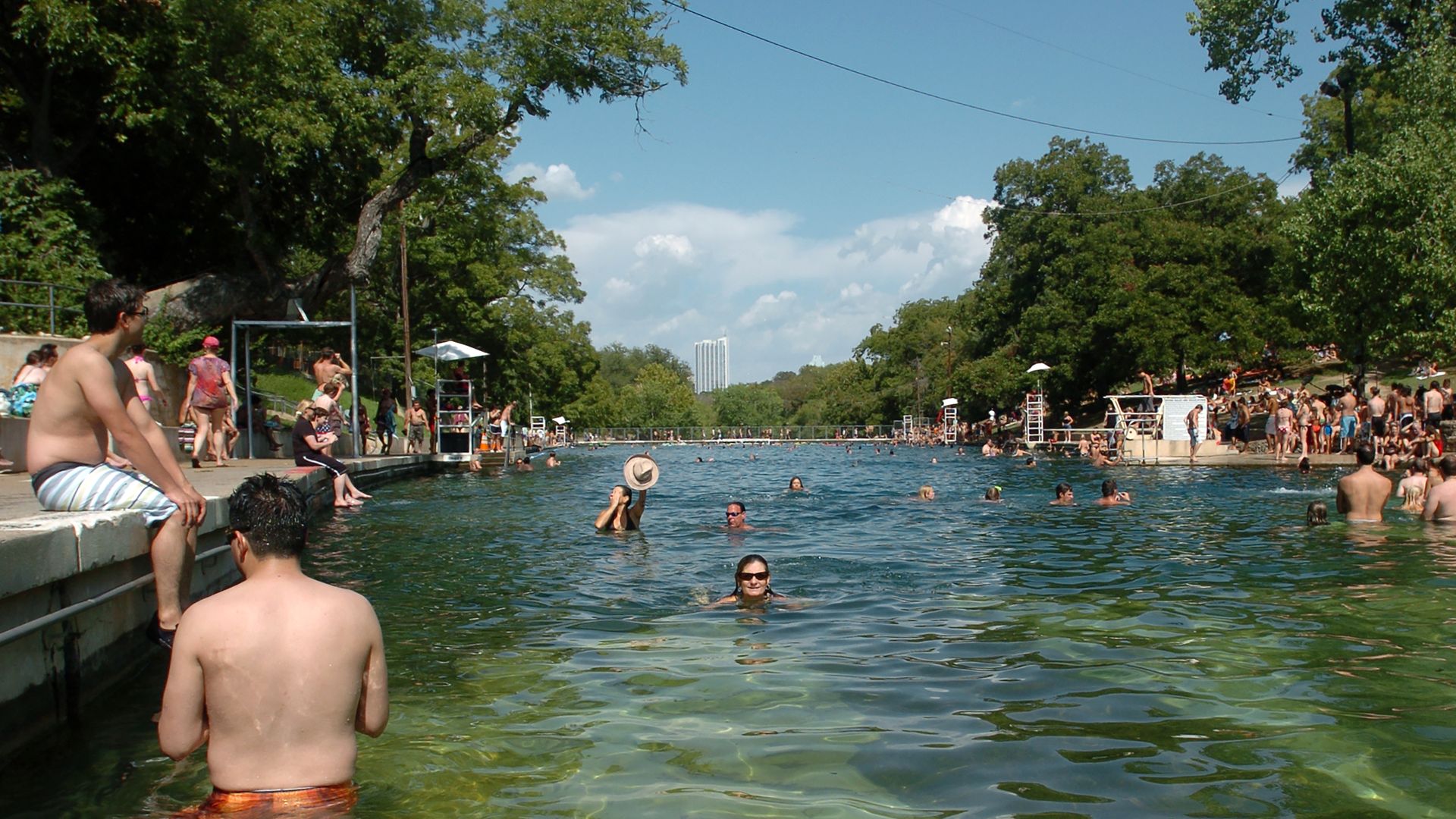 People swimming and sitting around a large outdoor pool with clear green-blue water, surrounded by trees and a blue sky with some clouds.