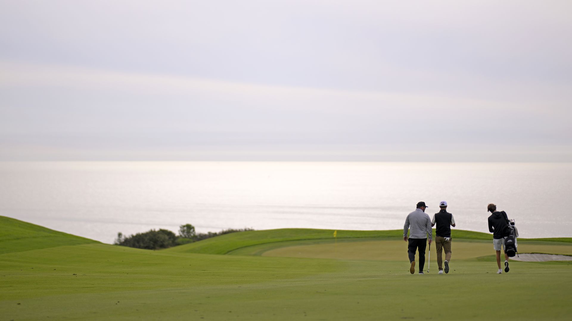A golfer, caddy and another man walk on the green fairway towards the hole at Torrey Pines Golf Course with a view of the Pacific Ocean. 