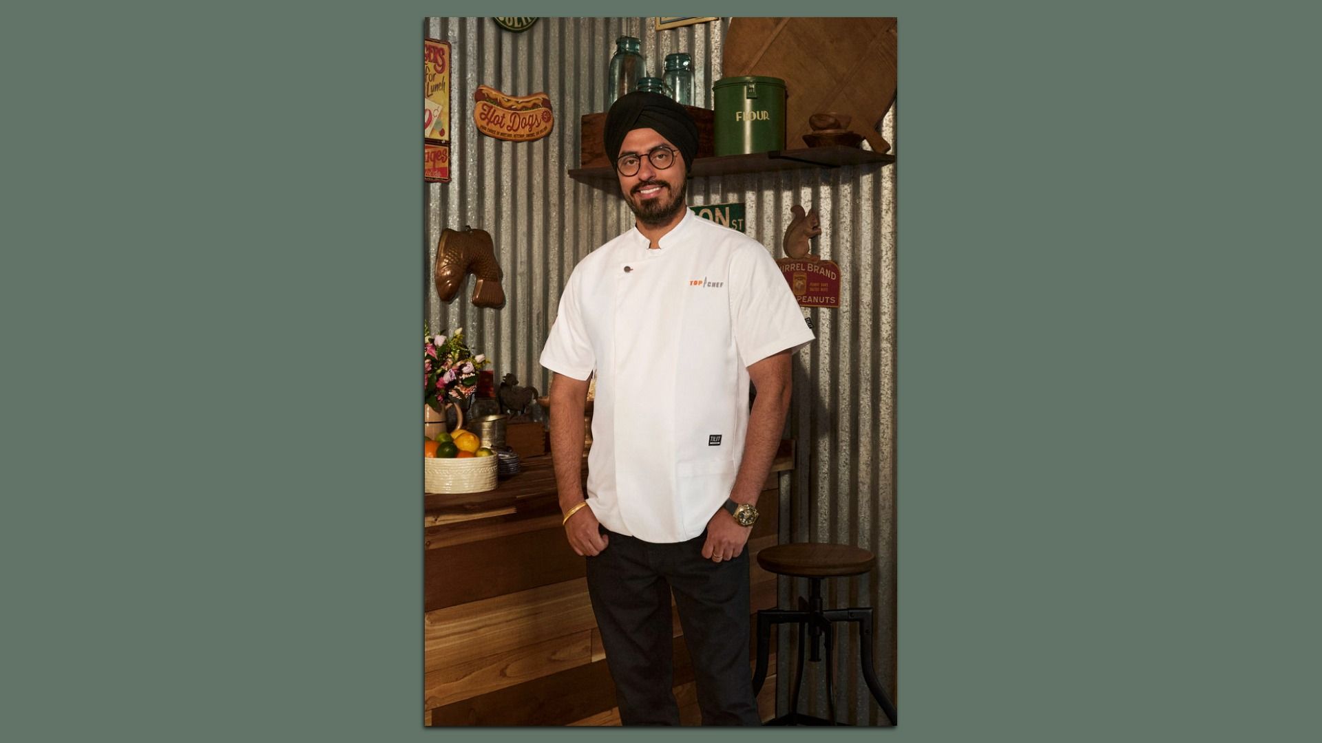 A smiling man wearing a black turban, glasses, and a white chef jacket with "Top Chef" logo stands in a rustic kitchen with metal walls, wooden shelves, and vintage signs.