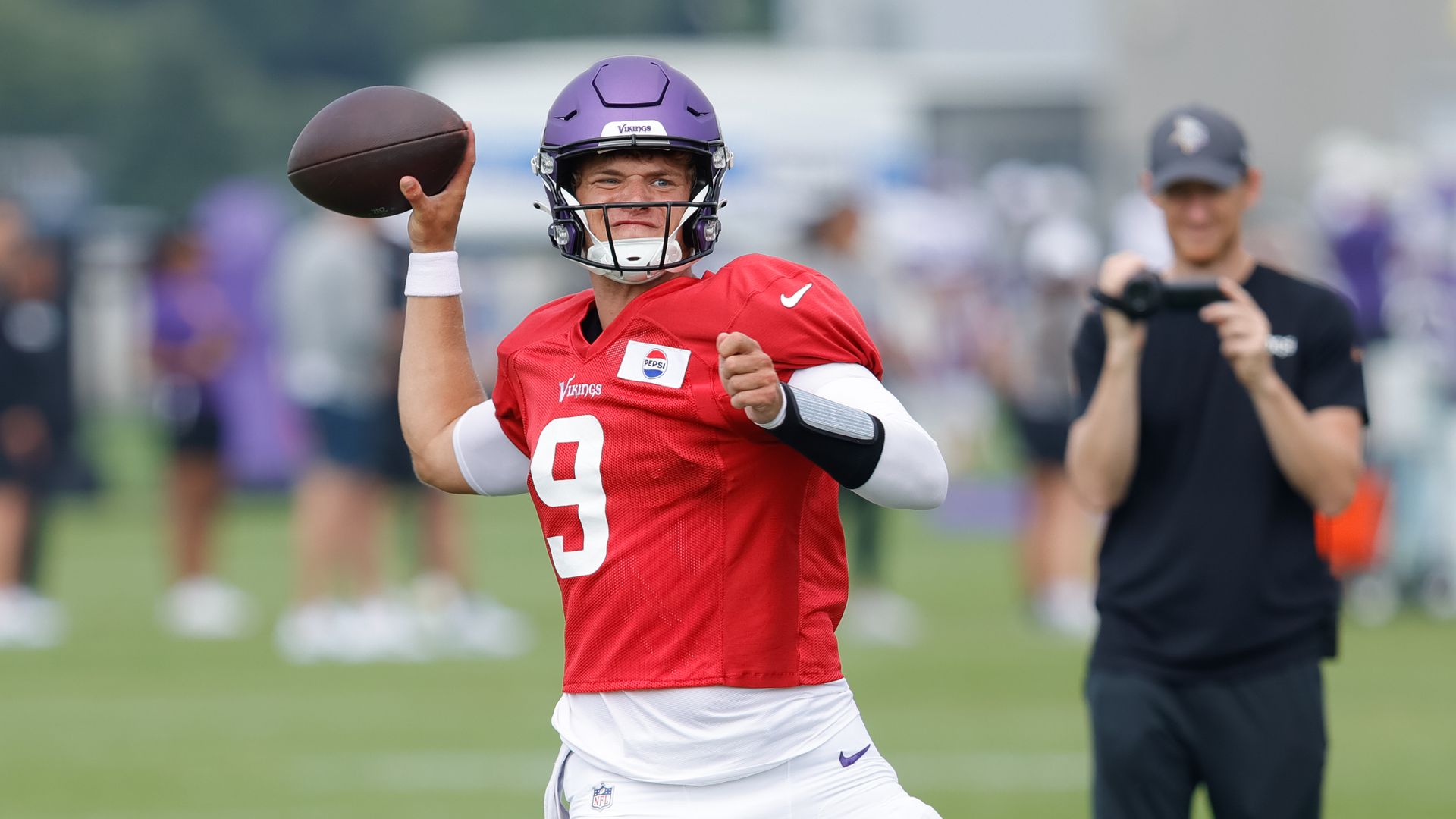 JJ McCarthy in a purple helmet and red jersey #9 preparing to throw a football on a green field during practice, with a person filming in black shirt and cap in background.