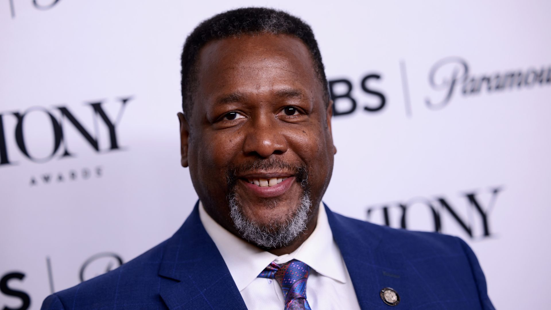 Photo shows Wendell Pierce smiling at the camera. He is wearing a blue suit jacket, white shirt and blue-patterned tie with a lapel pin.