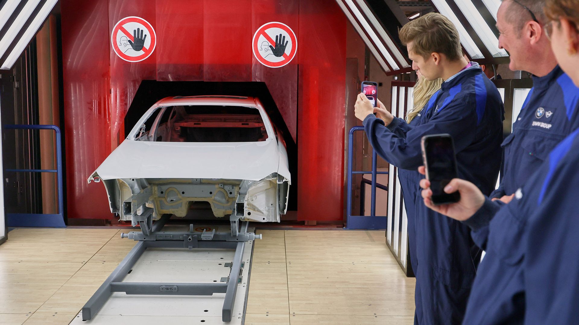 White car body on a gray frame in a red industrial chamber; workers in blue coveralls photograph the scene with smartphones as red no-hand safety signs hang overhead.