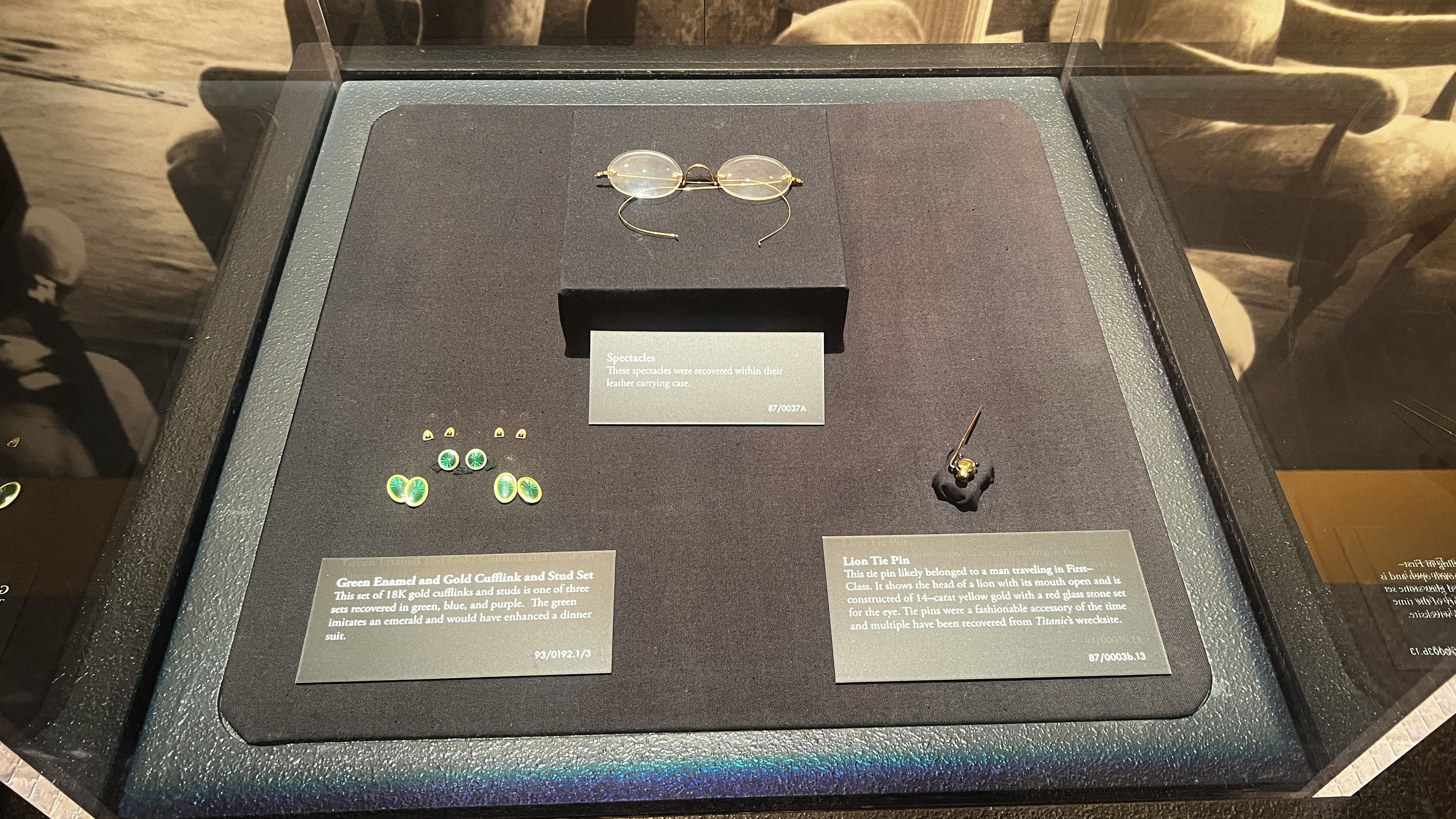 Display case of vintage accessories: center gold-rimmed round glasses on a black stand; left green and gold cufflinks; right a small golden pin. A placard reads that the spectacles were recovered.
