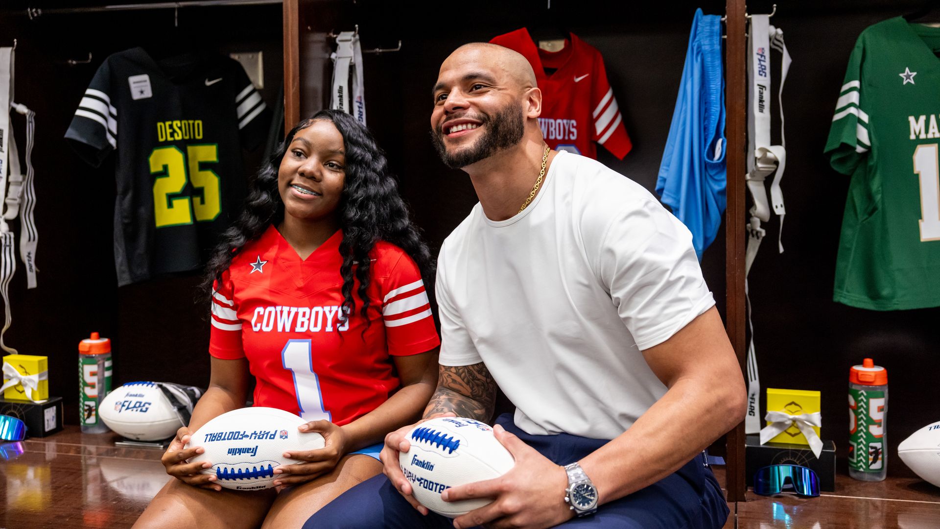 An athlete in a red uniform that says "Cowboys" sits next to a player, both holding footballs
