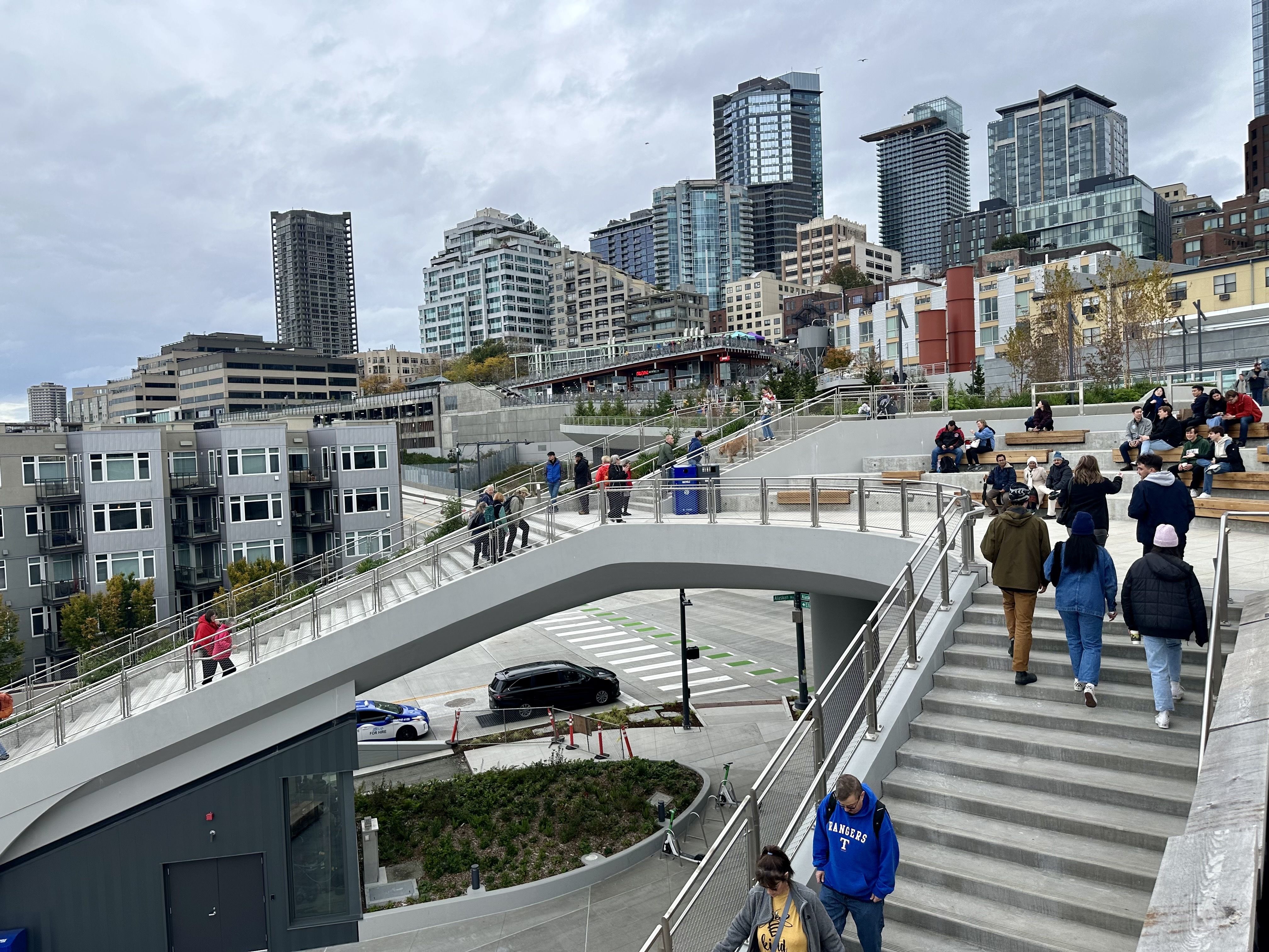 People walk up steps of stairs toward a terrace with skyscrapers in the background.