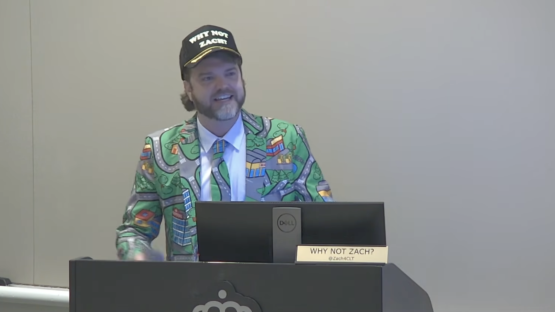 Man giving a talk at a podium in a bright map-pattern blazer and blue tie, wearing a black cap that reads "WHY NOT ZACH?"; nameplate on the desk says "WHY NOT ZACH? @Zach4CLT".