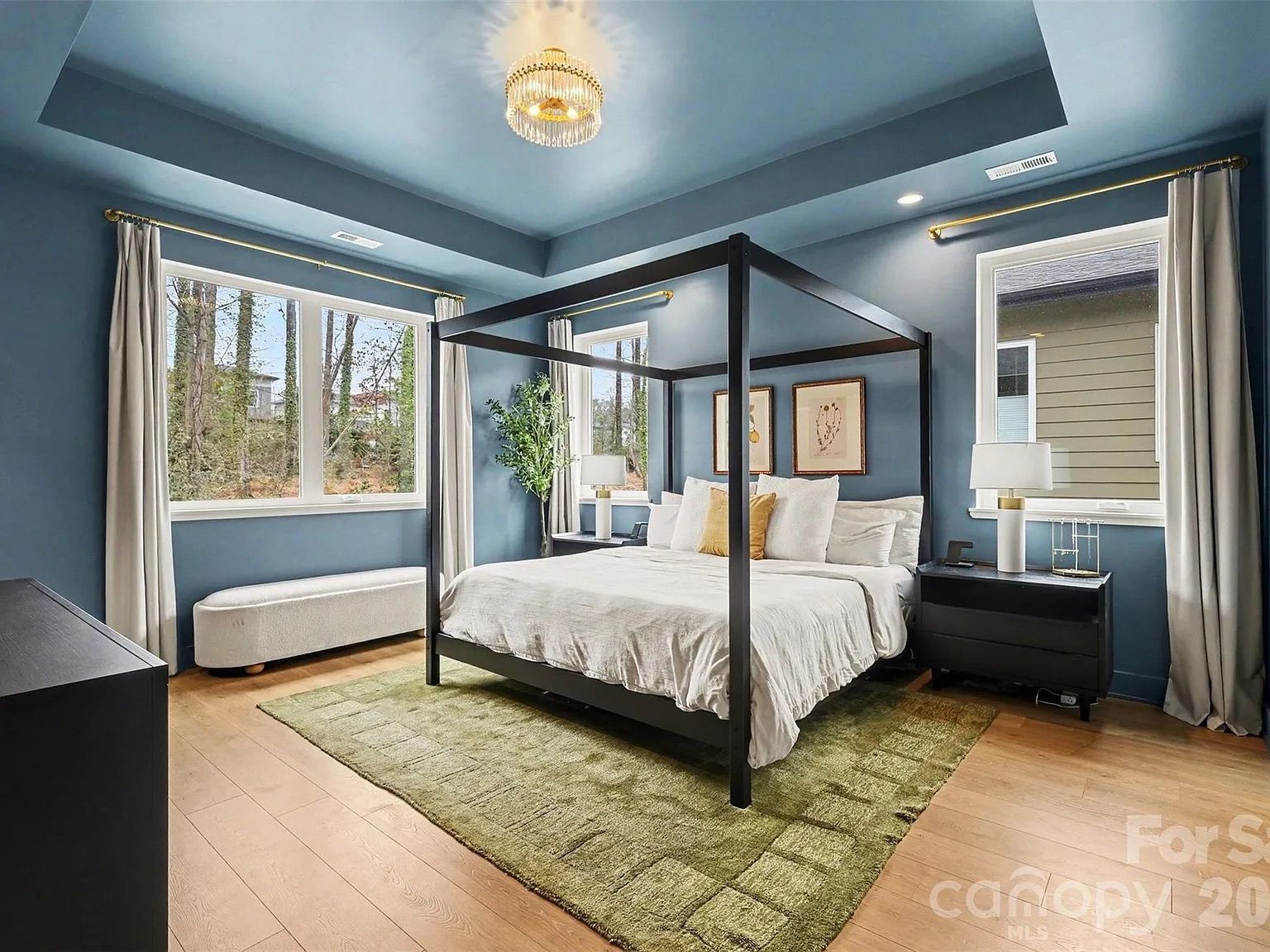Modern bedroom with blue walls and ceiling, black four-poster bed with white bedding and a yellow accent pillow, green rug, large windows with white curtains, and wood flooring.