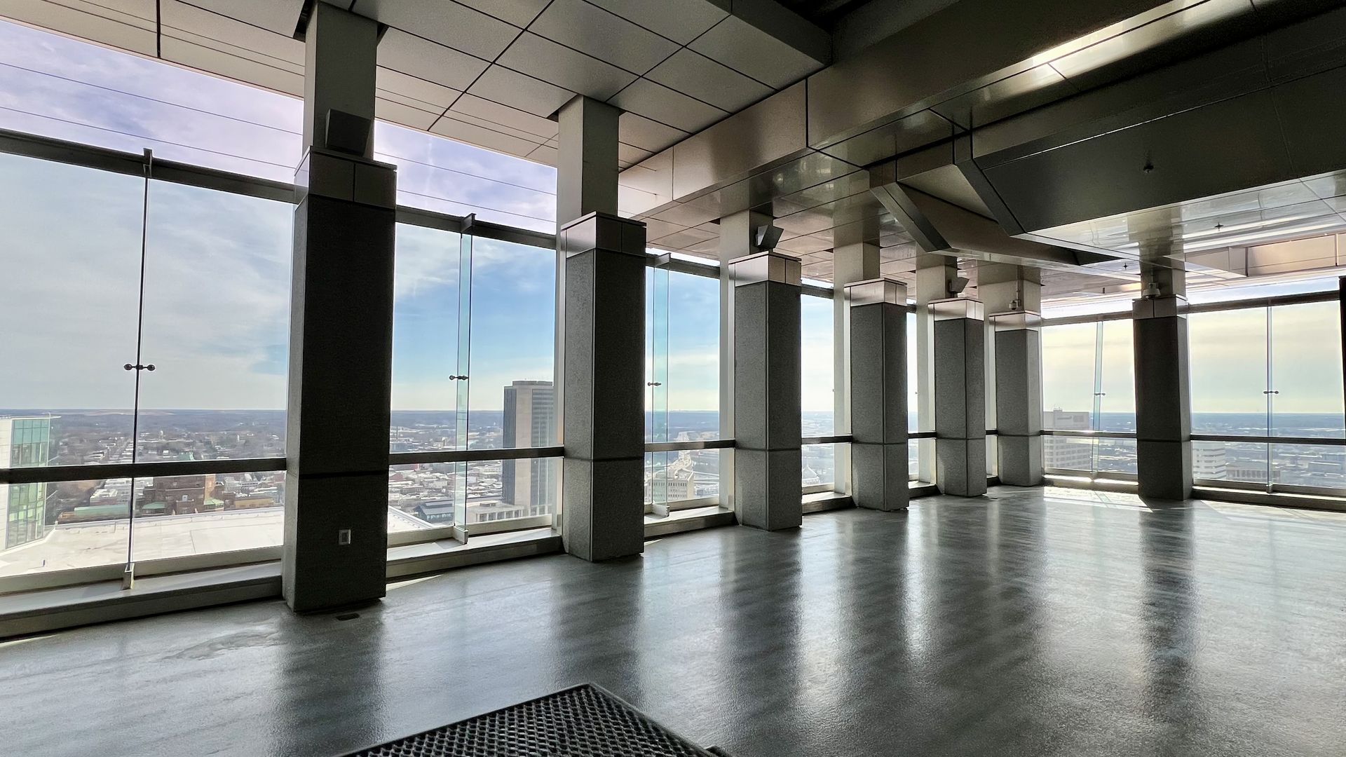 Empty indoor space with large floor-to-ceiling windows and multiple square pillars, offering a view of a cityscape under a partly cloudy sky during daytime.