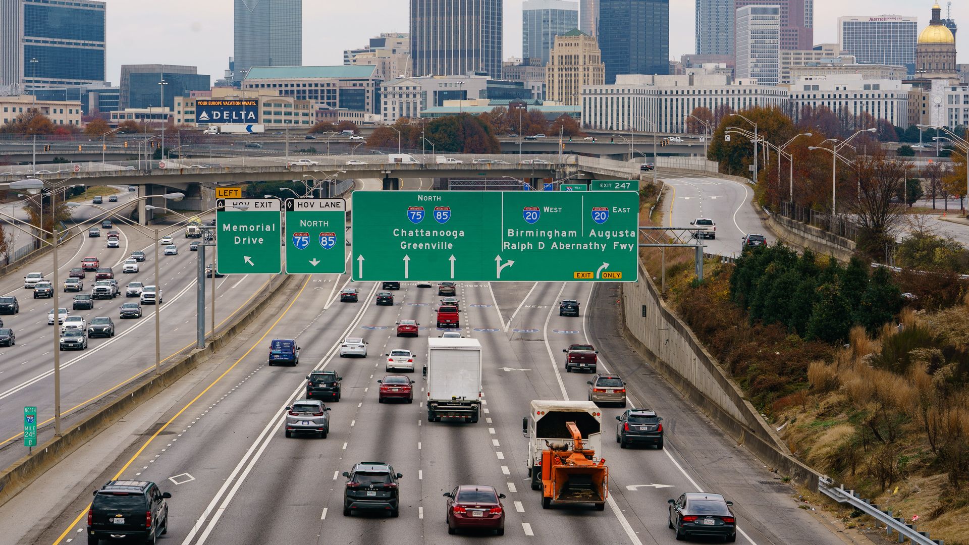 An aerial view of the northbound side of Interstate 75/85 at Interstate 20 in Atlanta. Vehicles are seen traveling along the interstate with trees, billboards and buildings in the background.