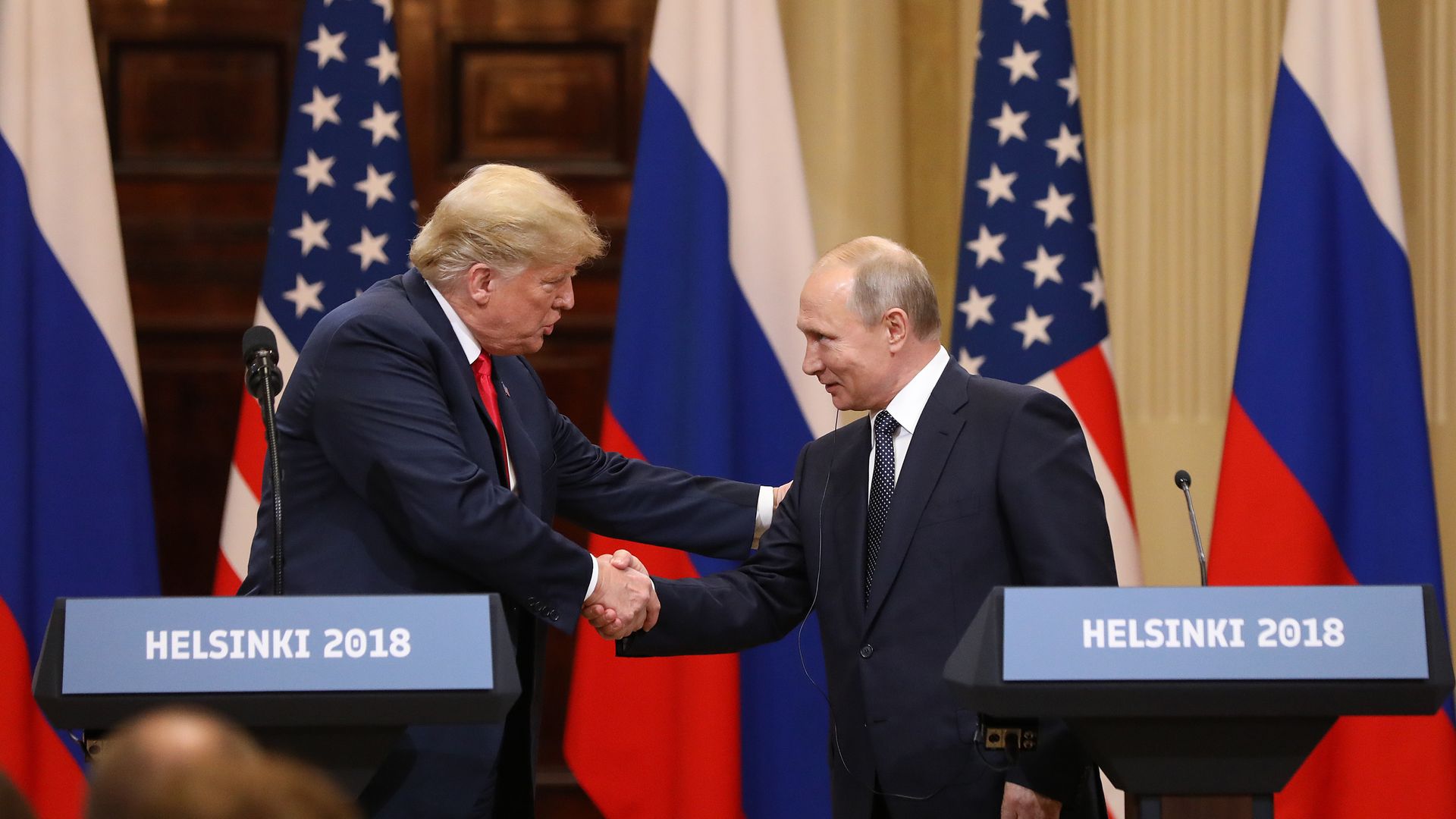 Donald Trump and Vladimir Putin shake hands in front of U.S. and Russian flags at podiums labeled "HELSINKI 2018" during a formal diplomatic event.