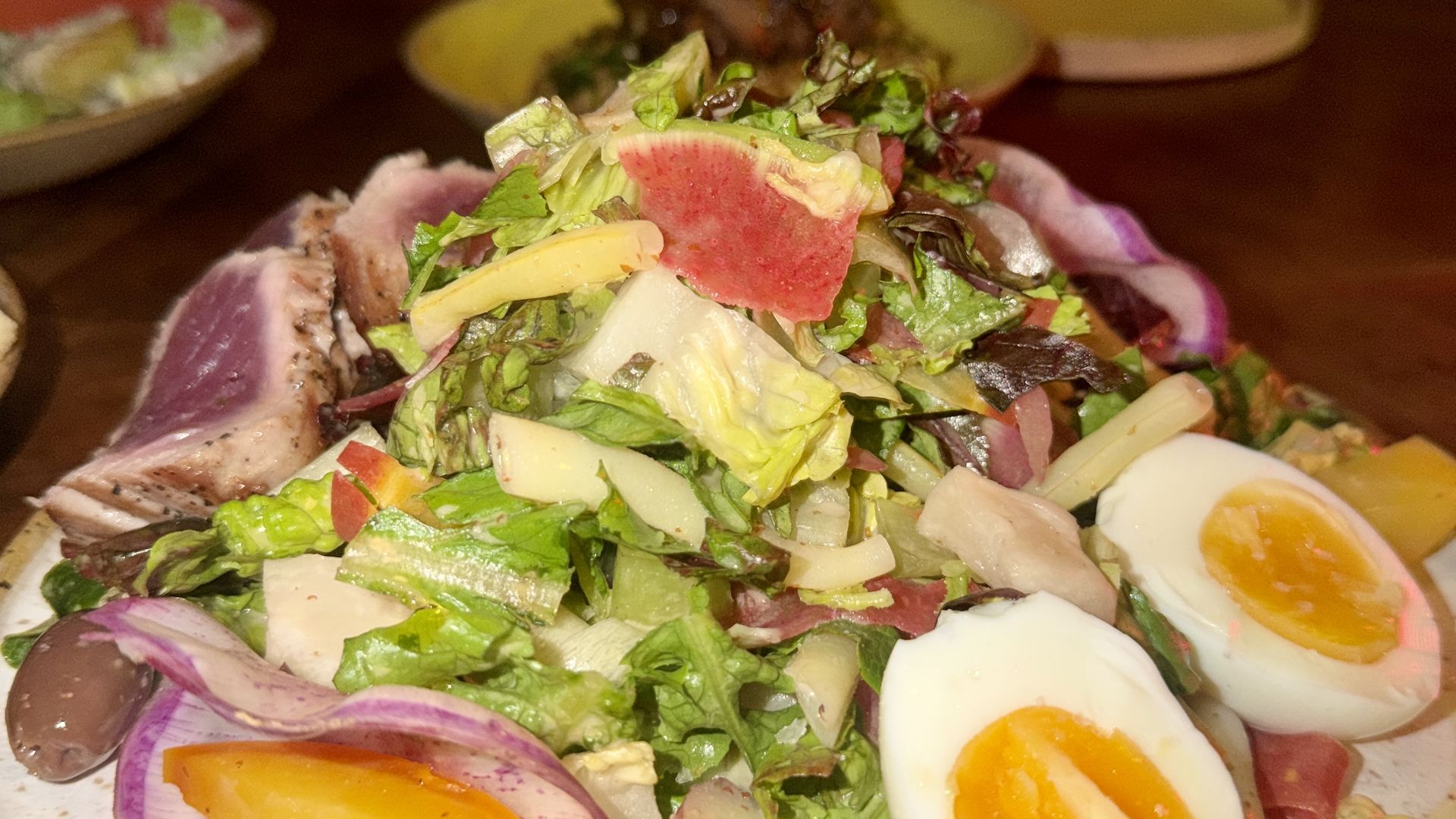 Close-up of a colorful salad on a plate: greens, radish, red onion, olives, purple potatoes, and halved soft-boiled eggs with orange yolks; two people blurred in the background.