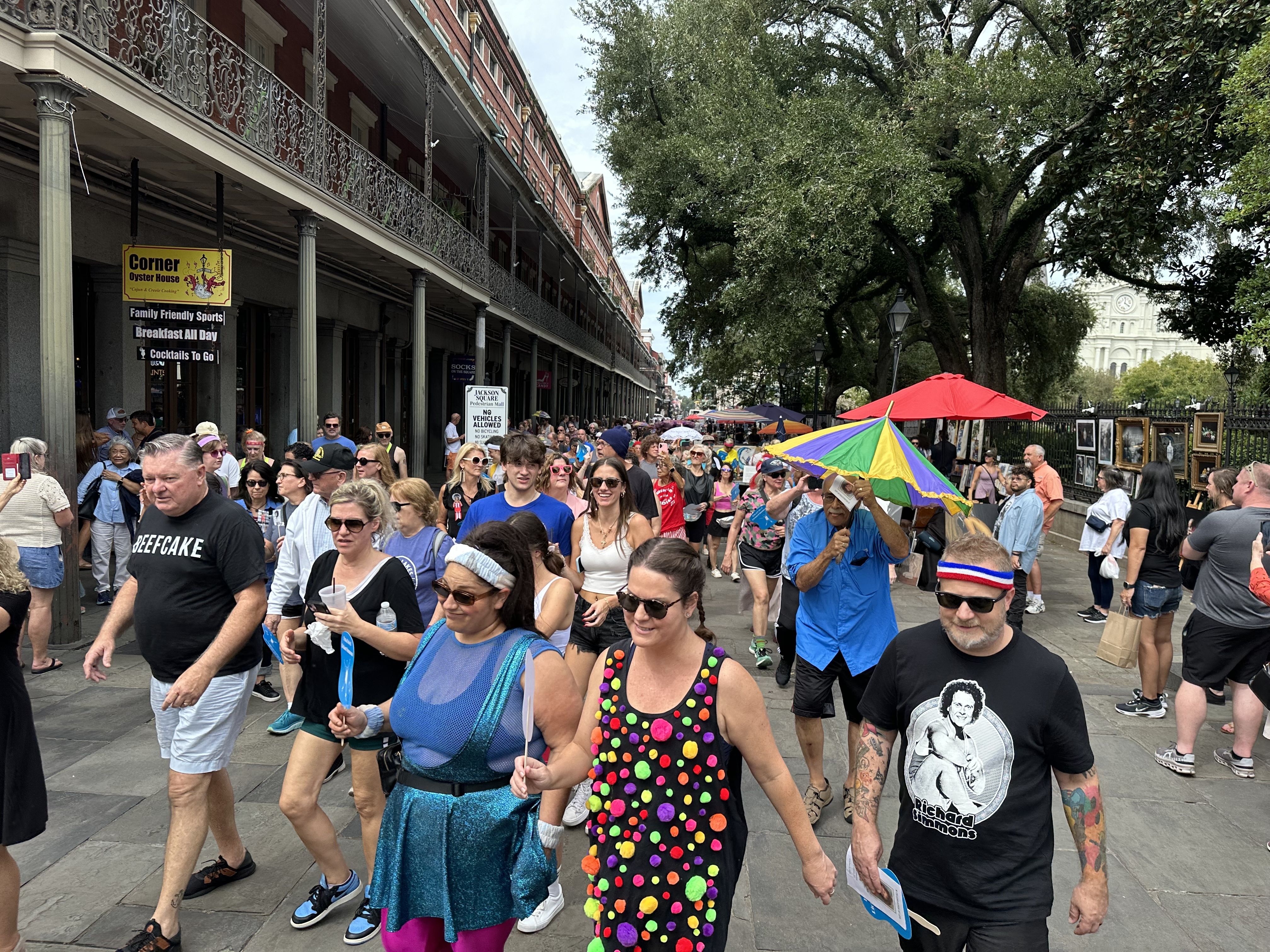 People wearing Richard Simmons T-shirts and 1980s-style workout gear and wigs walk past the camera.