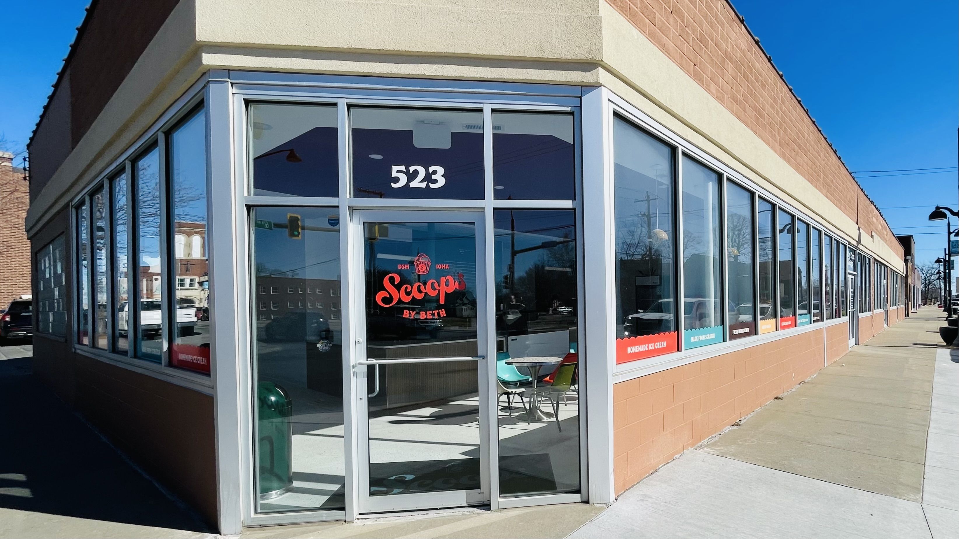 Corner ice cream shop with glass windows and door labeled 523 and red "Scoops by Beth" logo, colorful chairs visible inside, and clear blue sky outside.