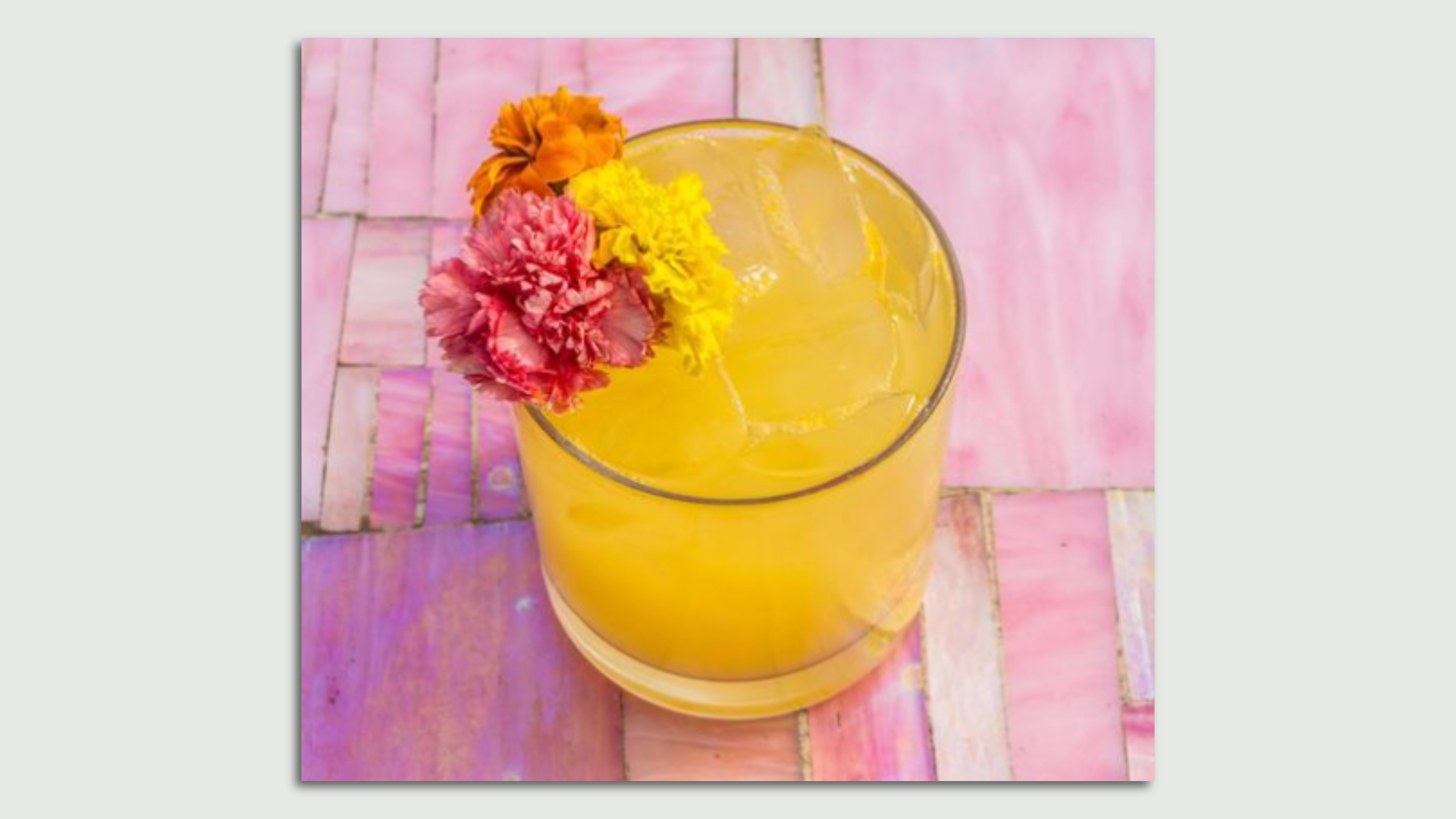 A yellow cocktail on a pink table with a pink flower on the rim.