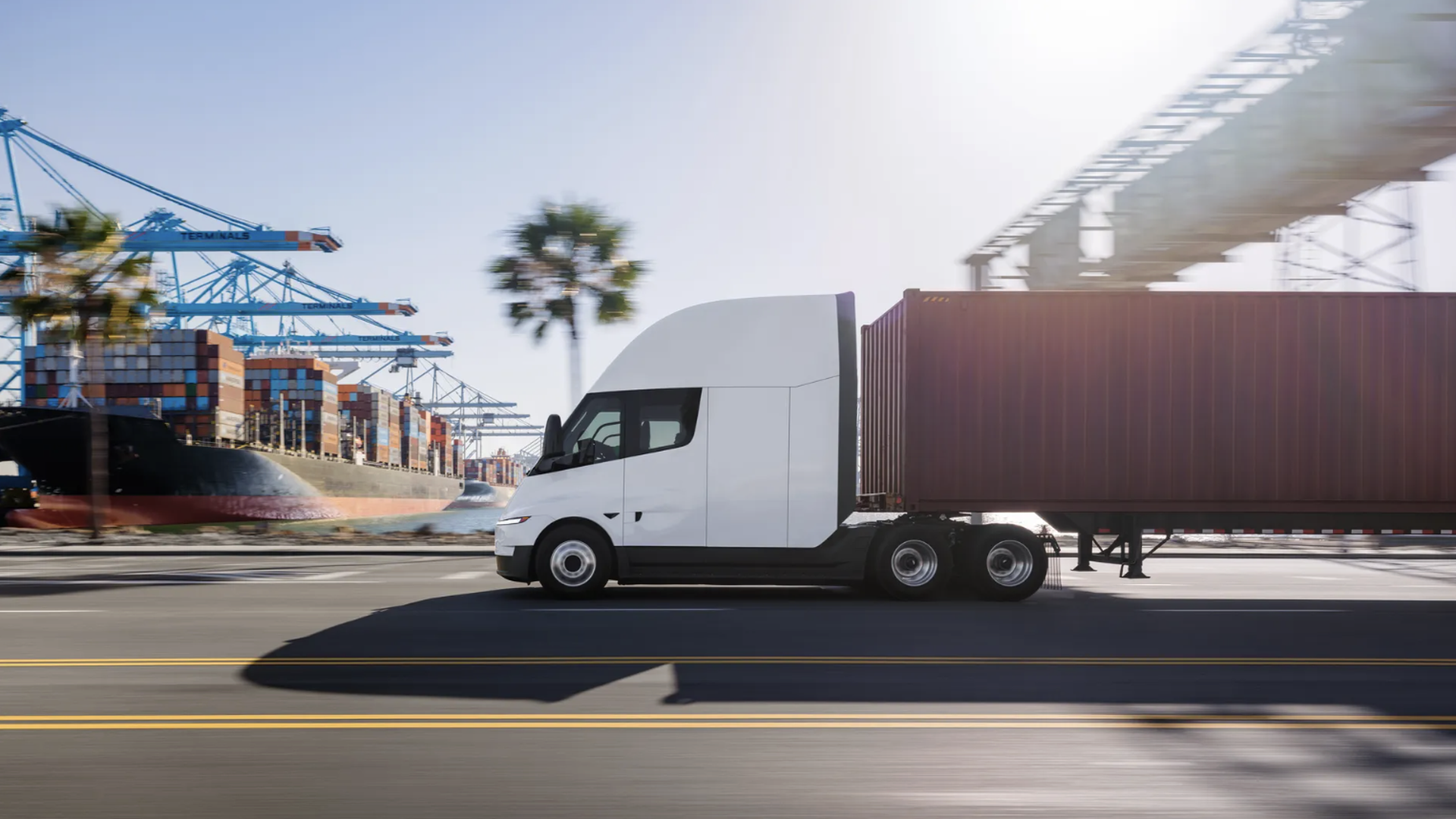 White semi-truck with a long brown shipping container trailer drives along a road beside a busy port, featuring blue gantry cranes, stacked colorful containers, a large ship, and palm trees.