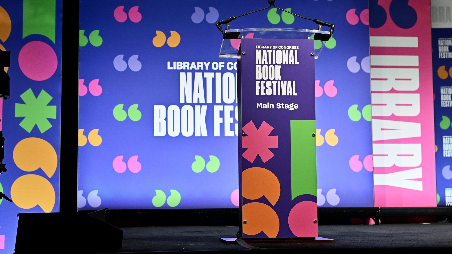 Podium on a bright festival stage at the Library of Congress National Book Festival; purple panel reads "Main Stage". Colorful backdrop with blue panels and scattered pink, green, orange quotation marks.