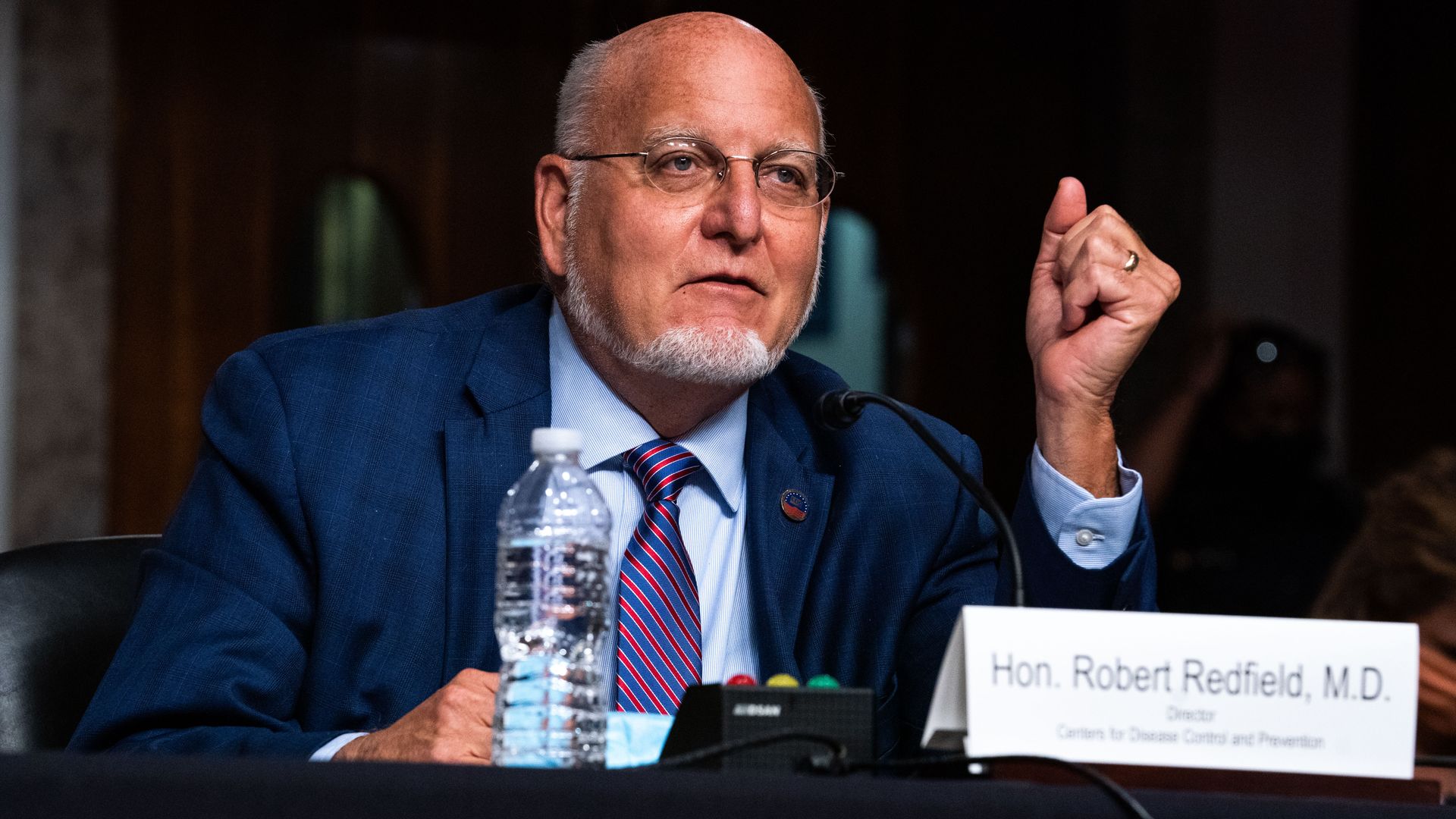Redfield wears a suit and tie while sitting in front of a name tag 