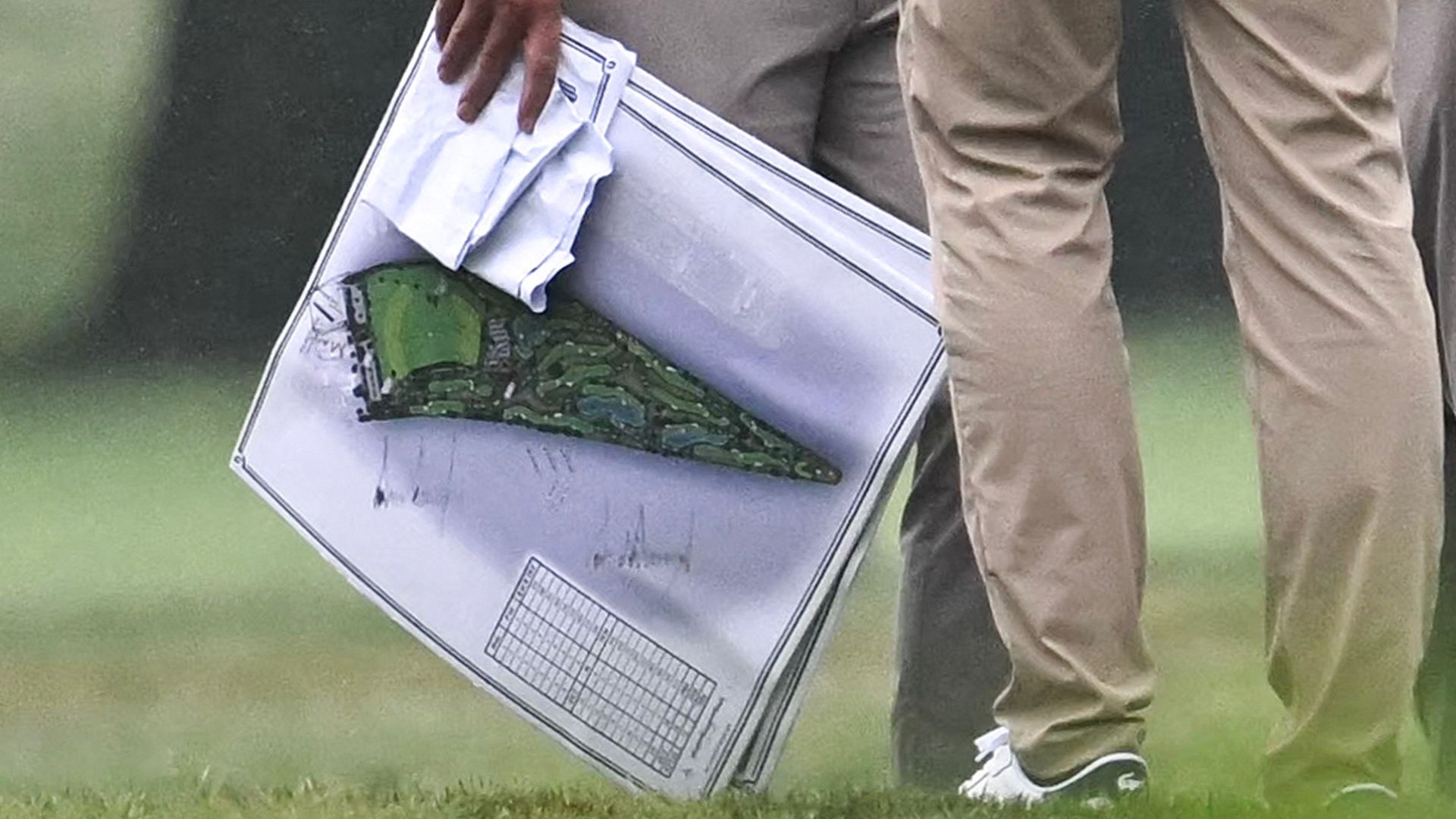 Person in tan pants and white sneakers stands on grass, gripping a large silver rectangular case opened at an angle to reveal a green camouflage interior and printed grids inside.