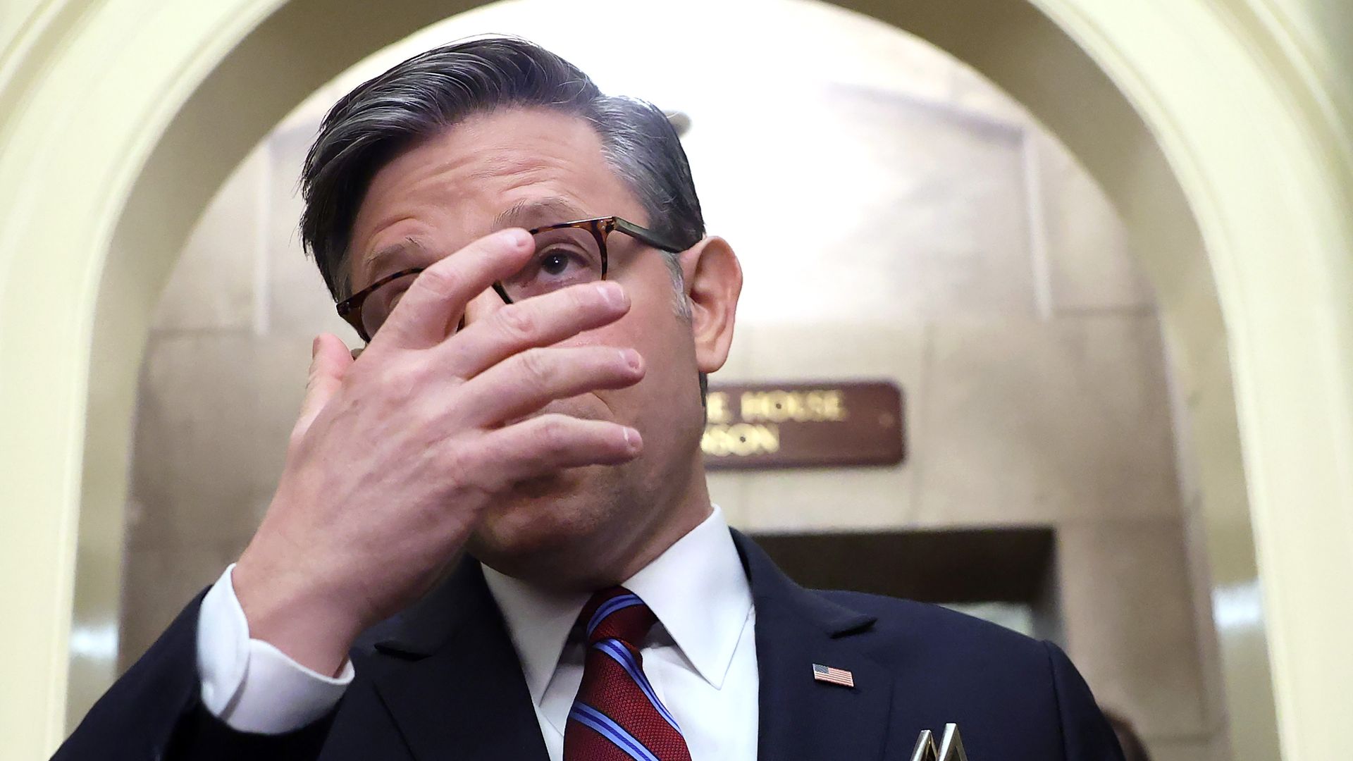 A man in a dark suit and red striped tie adjusts his tortoiseshell glasses with one hand, standing in a pale arched hallway, wearing a small American flag lapel pin.