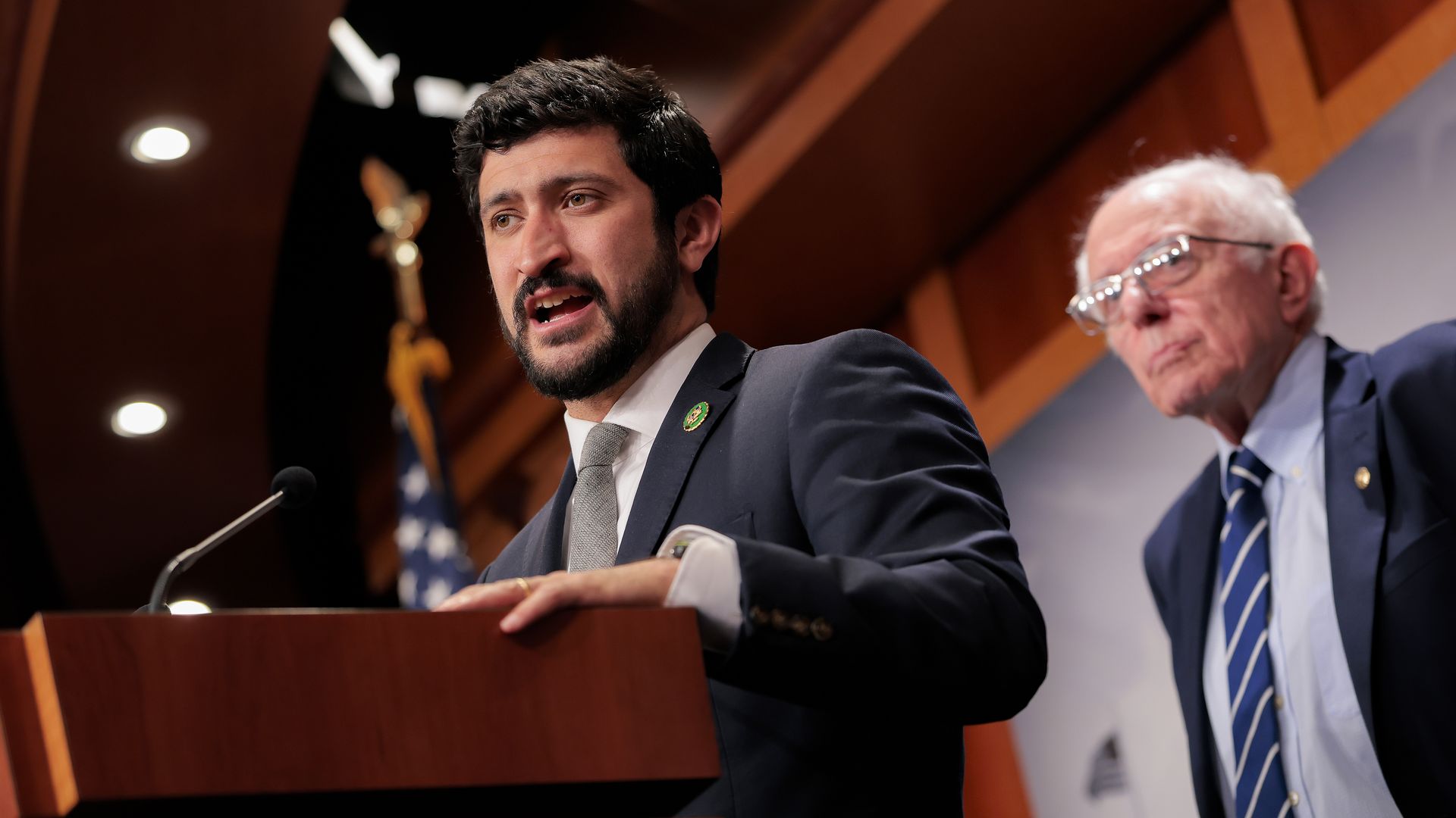 Two men in suits at a podium; the foreground speaker with dark hair and beard addresses a microphone, while another man with glasses stands beside him at a formal event.