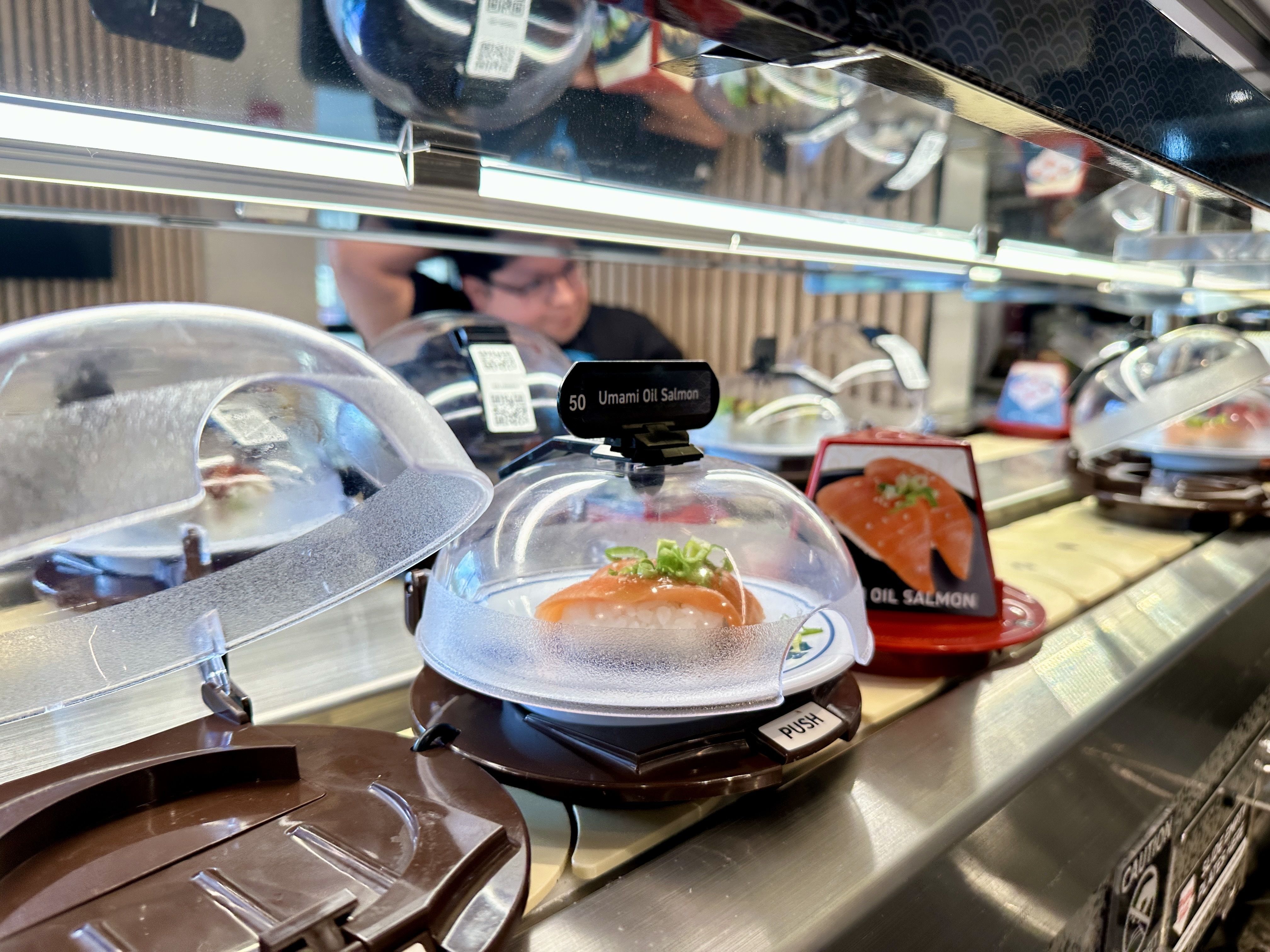 Sushi counter with glass domed lids over plated salmon, shiny metal rails, and a blurred staff member in the background; a label reads Umami Oil Salmon.