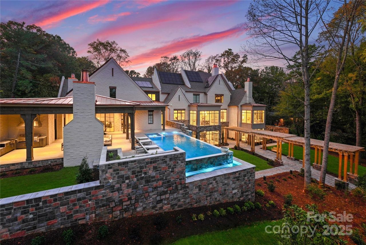 Luxurious large white house with lit windows at sunset, featuring an infinity pool with blue lighting, stone walls, wooden pergola, and surrounding trees and yard.