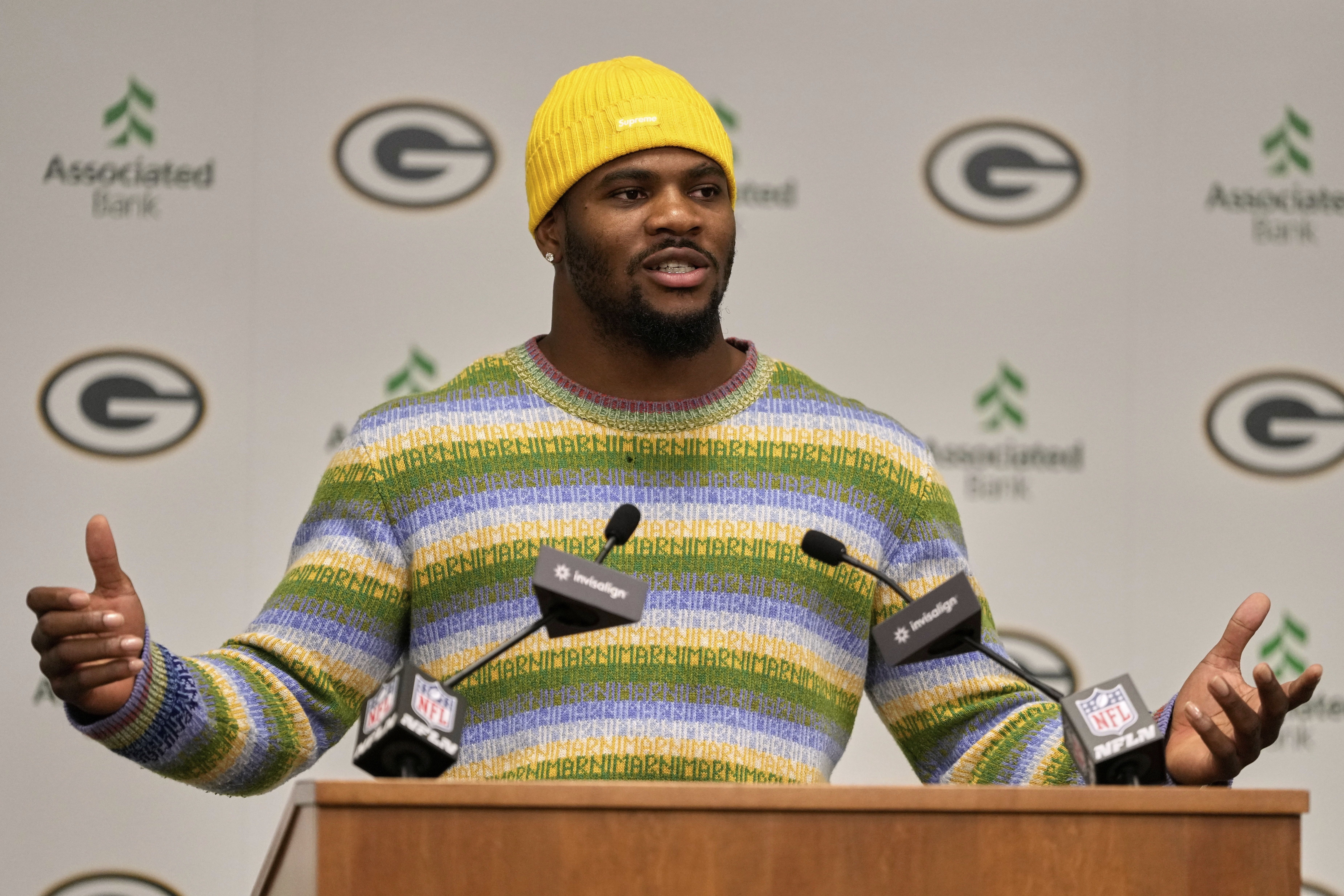 Man wearing a bright yellow beanie and a colorful striped sweater speaks at a podium with microphones, with Green Bay Packers logos and Associated Bank logos on the backdrop.