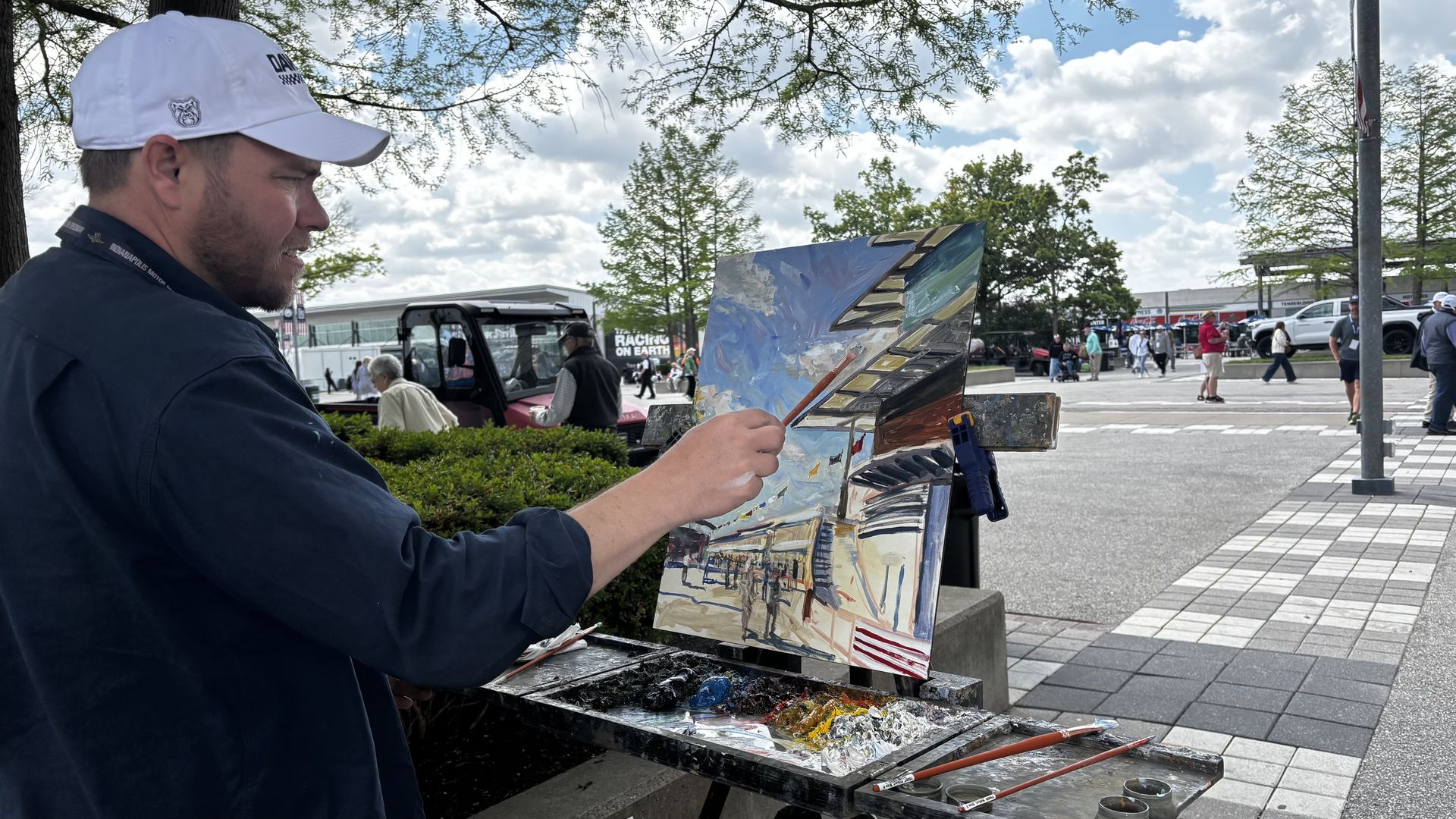A man in a white cap and dark jacket paints on a canvas mounted on a portable easel outdoors, in a sunny plaza with trees, clouds, pedestrians, and parked cars.