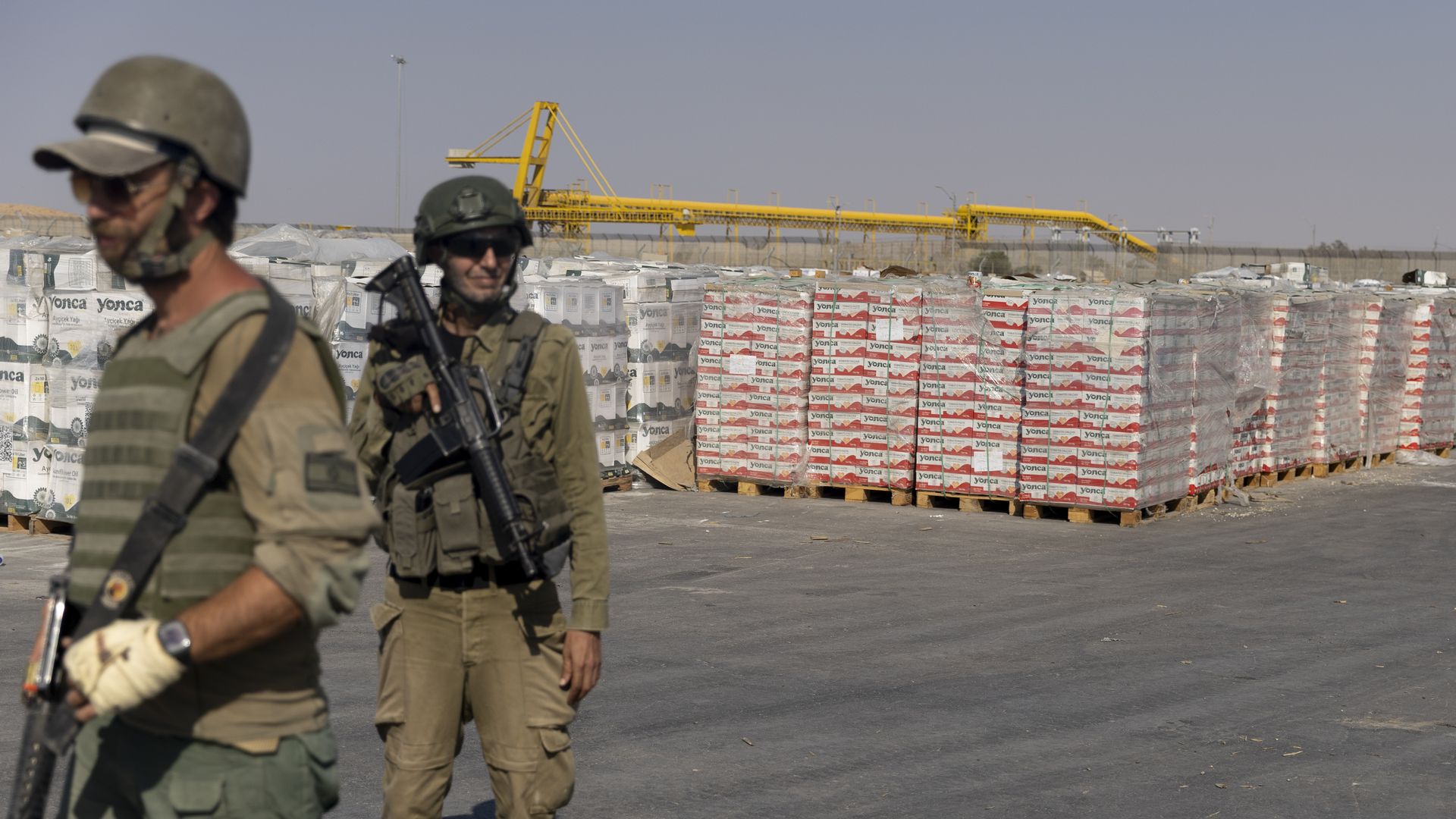 Two armed soldiers in uniform and helmets standing near large stacks of packaged goods wrapped in plastic on pallets in an industrial outdoor setting under a clear sky.