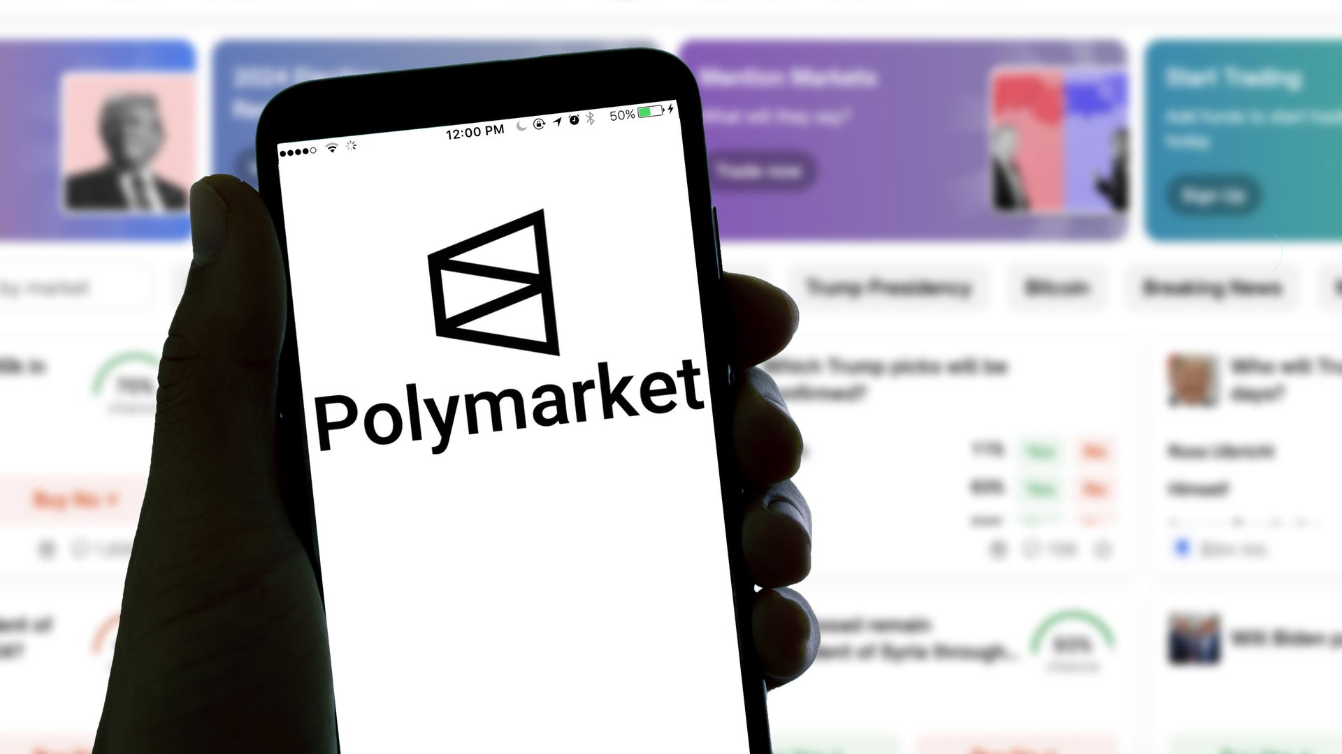 A hand holds a smartphone displaying the Polymarket logo; the blurred background shows a colorful website banner.